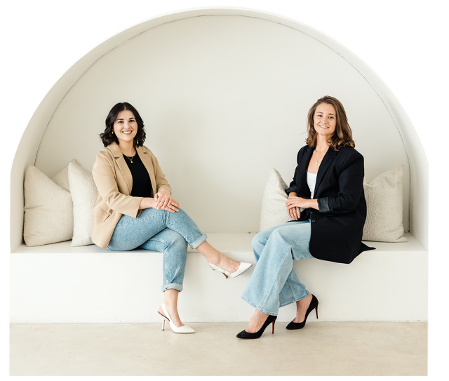 Two women (Sarah Clayton and Aly Nicolaou) sitting and smiling inside a decorative wall featuring a tropical palm tree design. .The setting has warm tones and a relaxed atmosphere