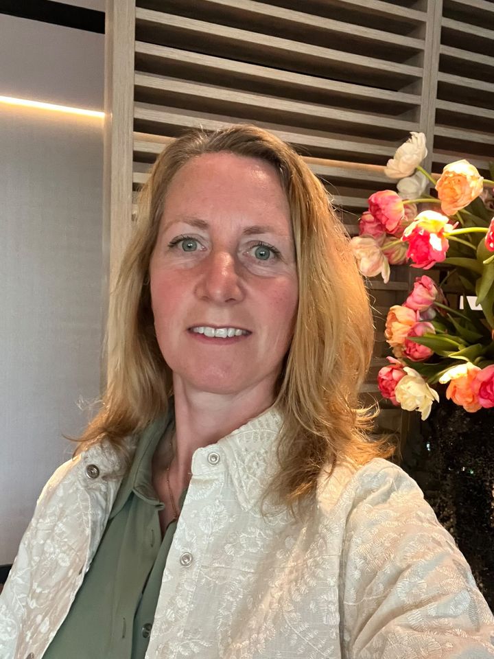 Paula in een groene blouse en lichte vest, glimlachend voor een houten wand met bloemen