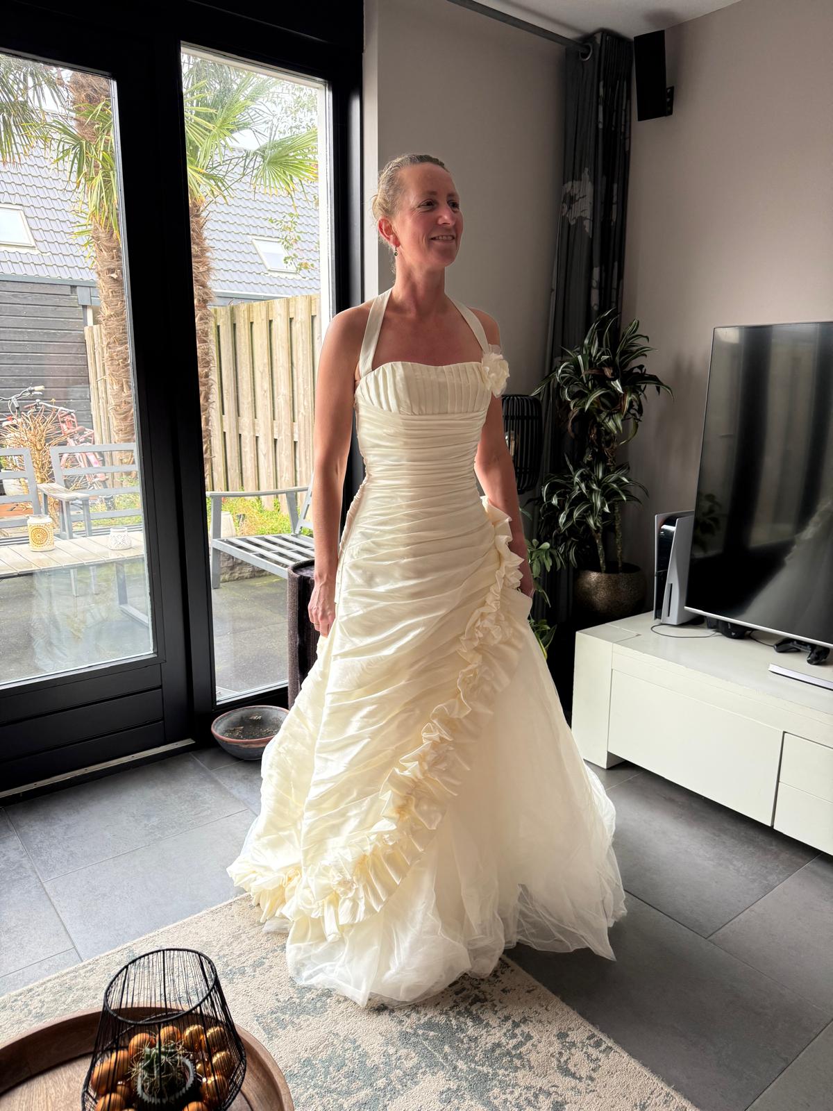 Een vrouw in een ivoorkleurige trouwjurk met plooien en ruches, haar haar opgestoken, in een woonkamer met een glazen deur naar de tuin
