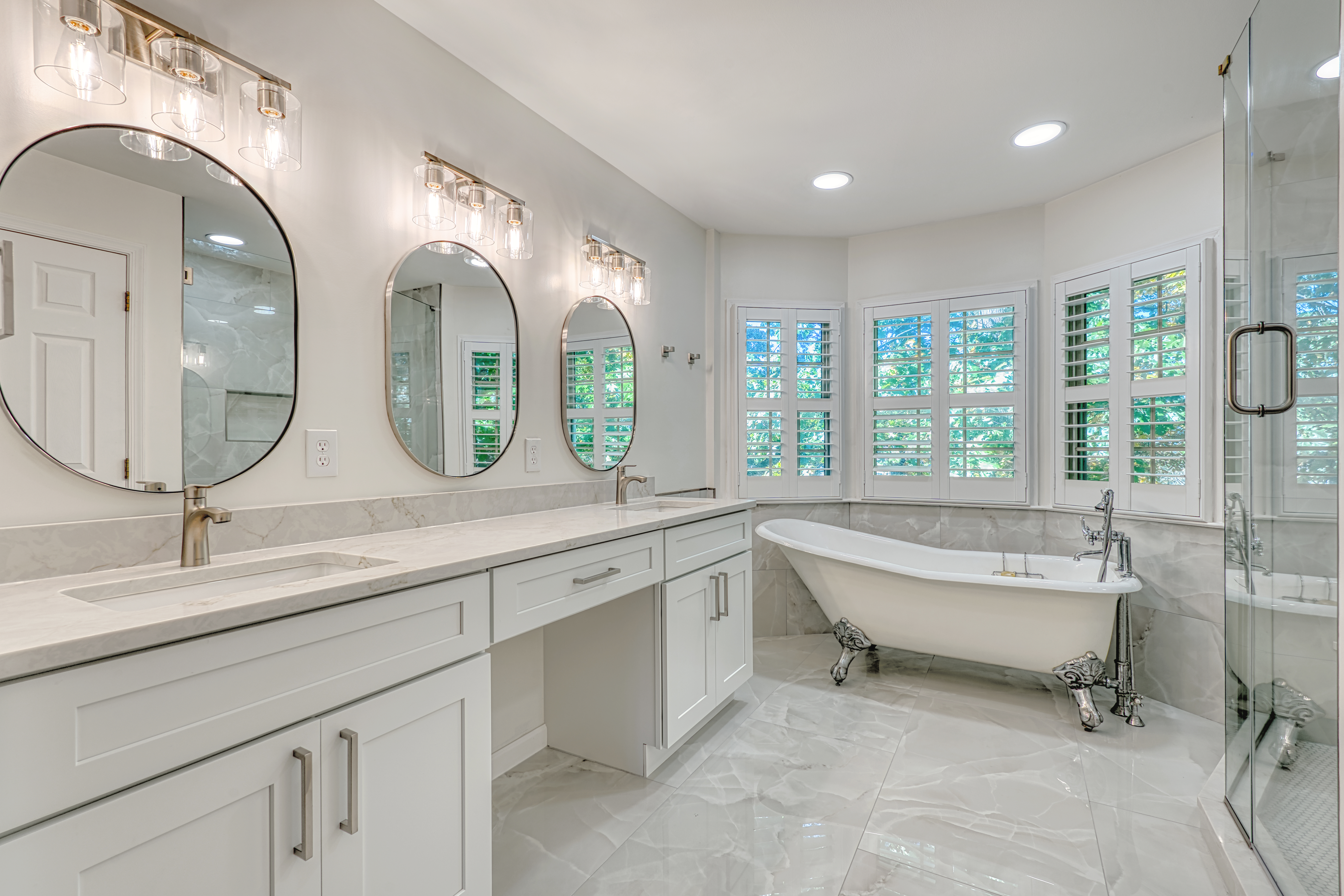 Luxury primary bathroom remodel in Chantilly, VA, featuring a double vanity with marble countertops, round mirrors, and a classic white clawfoot soaking tub by Reno Design Inc.