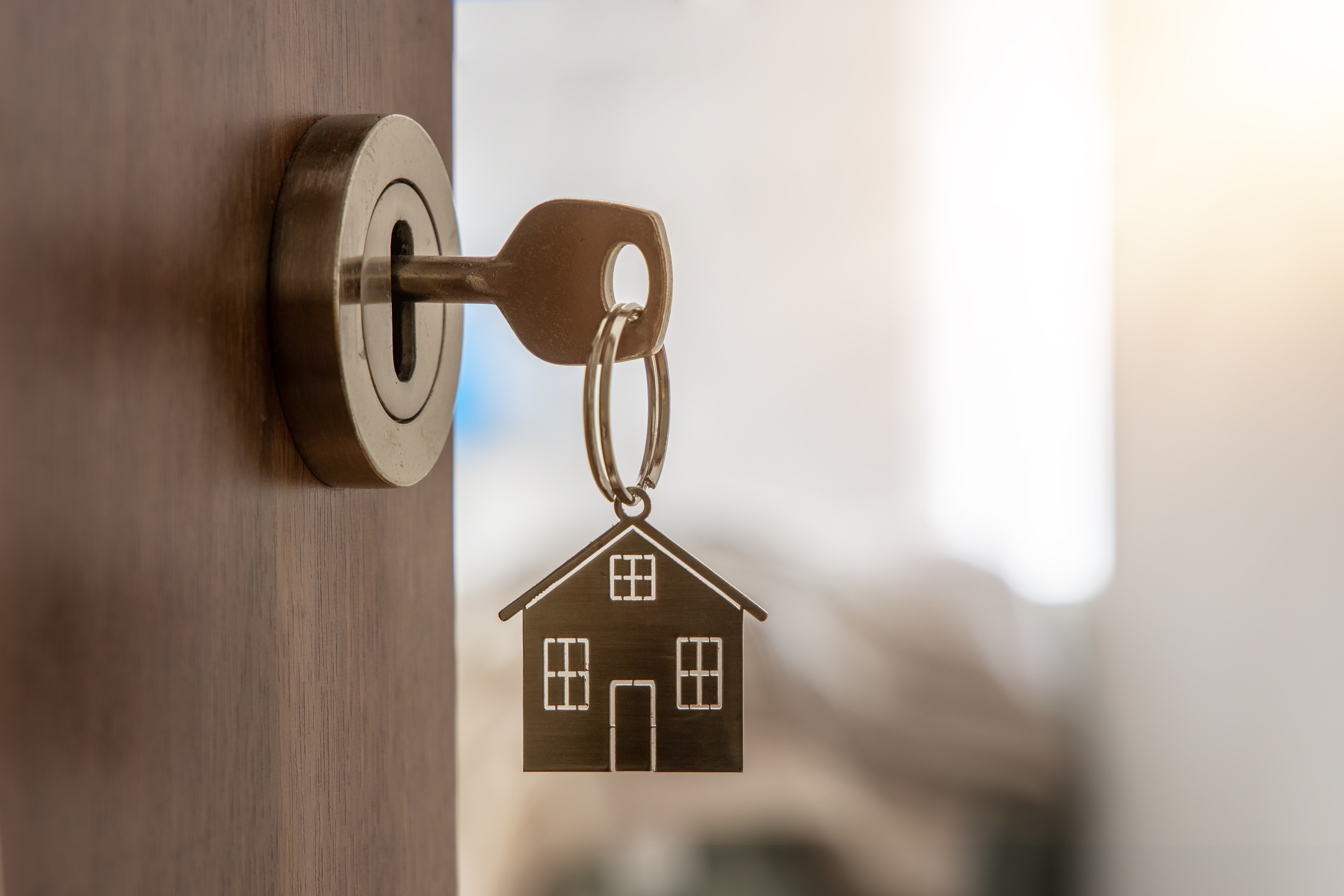 Close-up of a key inserted into a door lock, with a house-shaped keychain hanging from the key, representing housing and homeownership.