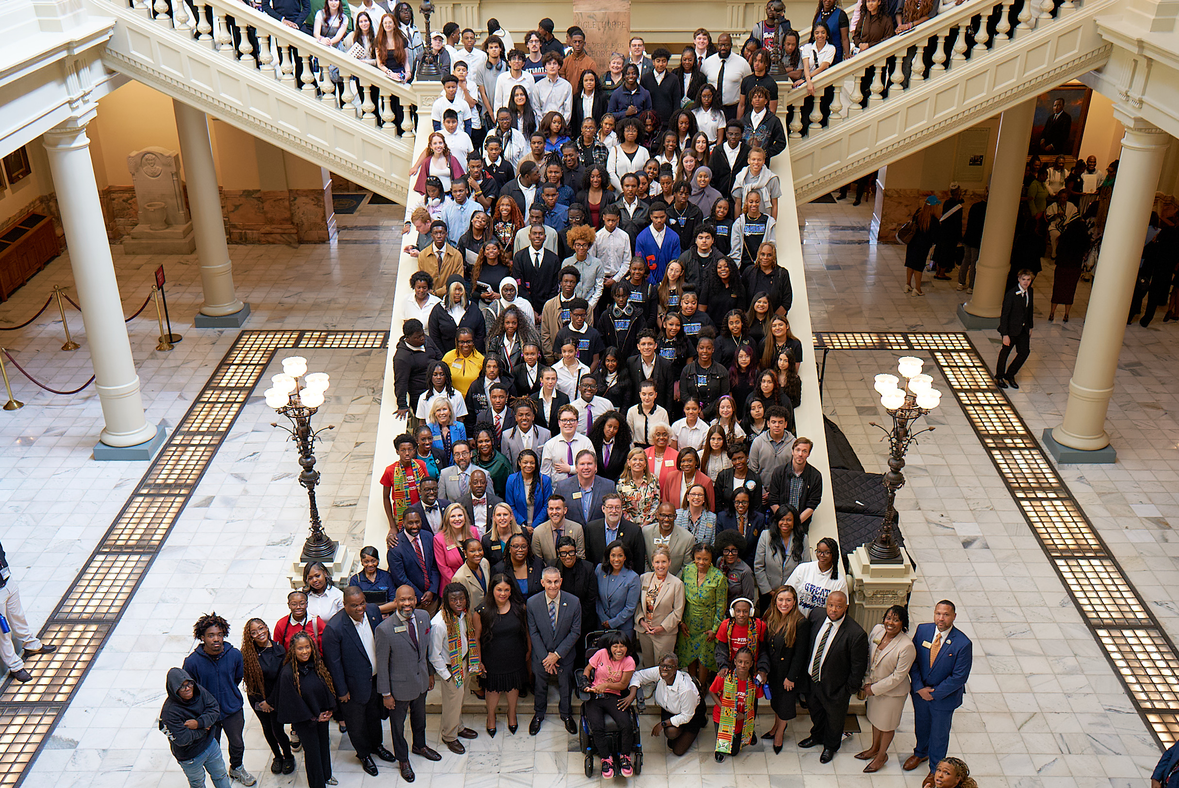 Large group photo at DeKalb Day at the Capitol showing dozens of students, community members, and local officials gathered on a grand staircase inside the Georgia State Capitol. The group fills the steps from top to bottom, with adults and youth standing closely together and facing the camera. At the front, several officials and organizers stand in a row, while additional attendees line both sides of the staircase and upper level. The setting features marble floors, ornate railings, and decorative lighting.