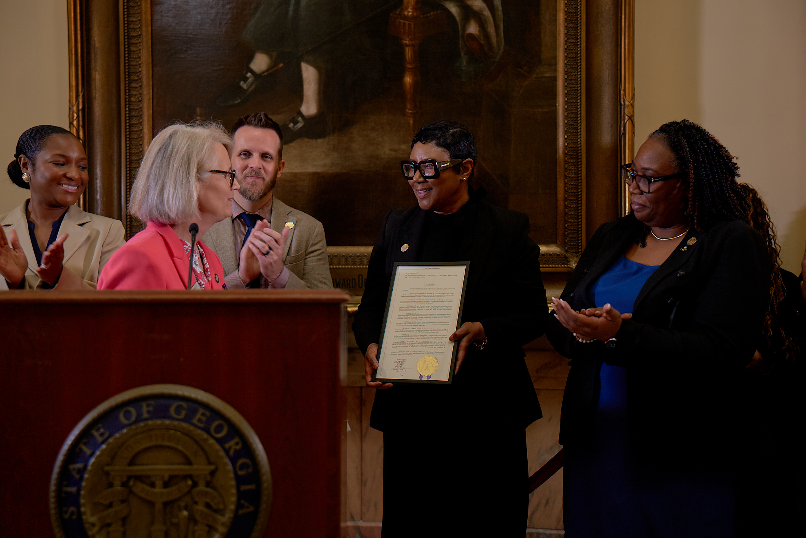 A woman in a pink blazer speaks at a podium with the State of Georgia seal during DeKalb Day at the Capitol. Behind her, Deputy Presiding Officer Dr. LaDena Bolton (Super District 7) and Commissioner Ted Terry (Super District 6) stand with DeKalb County CEO Lorraine Cochran-Johnson, who holds a framed proclamation. Commissioner Chakira Johnson (District 4) stands at right, applauding. The group is gathered in a formal government setting with a large portrait in the background.