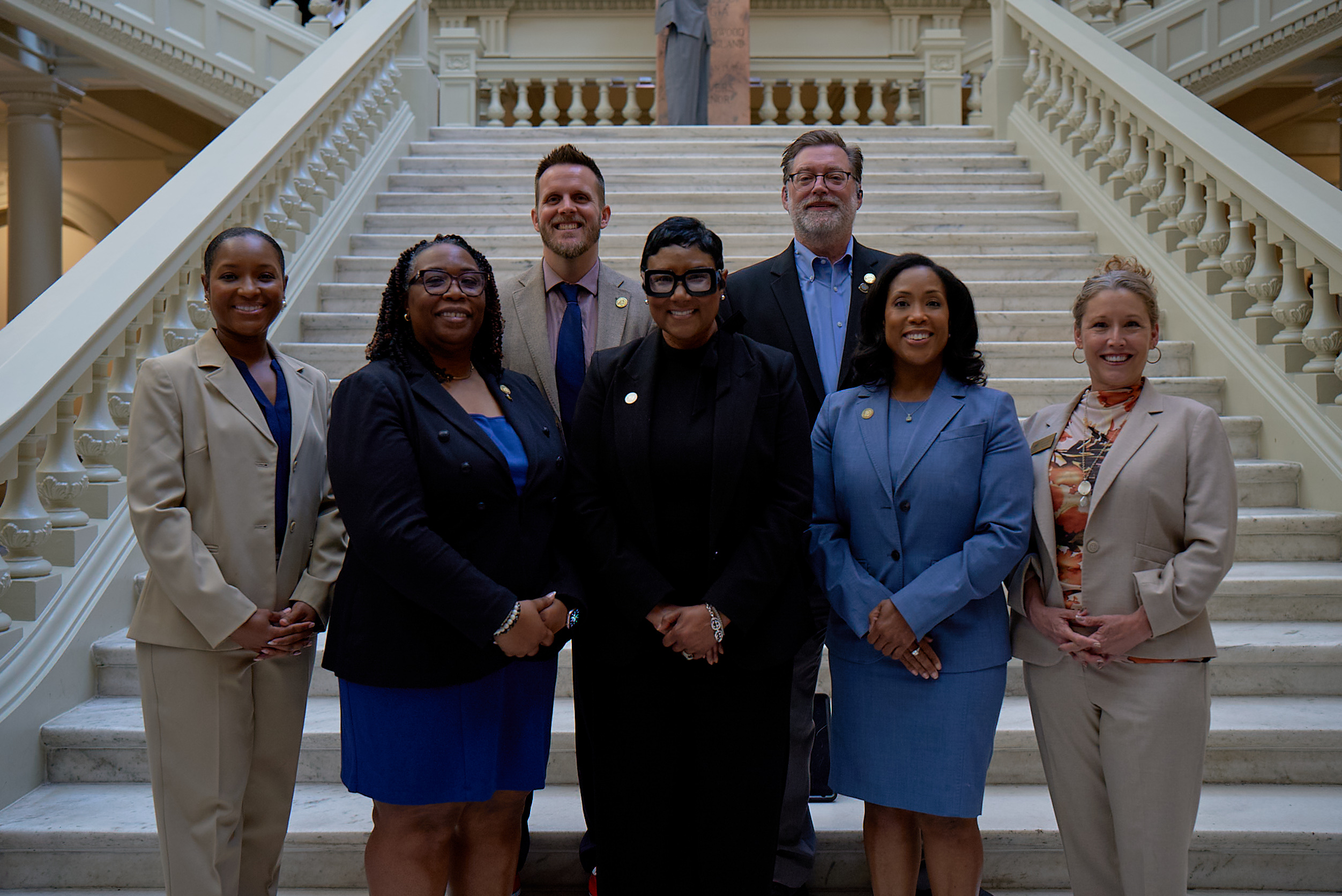 Group photo of the DeKalb County Board of Commissioners during DeKalb Day at the Capitol. Seven commissioners stand together on a grand staircase inside the Georgia State Capitol, smiling and facing the camera. Front row, left to right: Commissioner Dr. LaDena Bolton (Super District 7), Commissioner Chakira Johnson (District 4), DeKalb County CEO Lorraine Cochran-Johnson, Commissioner Nicole Massiah (District 3), and Commissioner Michelle Long Spears (District 2). Back row: Commissioner Ted Terry (Super District 6) and Commissioner Robert Patrick (District 1). The group is dressed in professional attire, posed in a formal government setting with white marble steps and ornate railings.