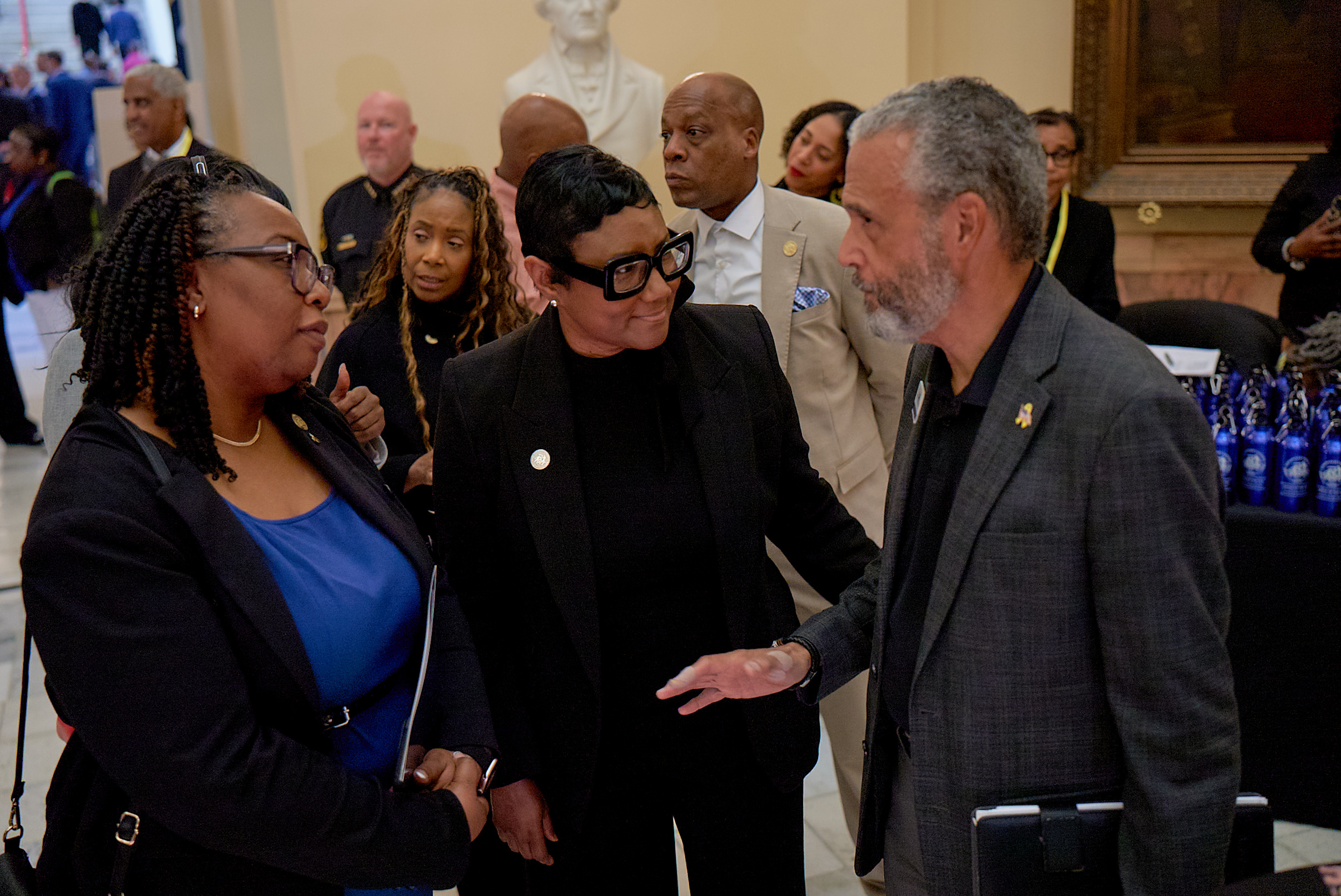 Commissioner Chakira Johnson and DeKalb County CEO Lorraine Cochran-Johnson speak with Rick E. Cunningham at an indoor event. Commissioner Johnson, wearing a blue top and dark blazer with glasses and braided hair, stands on the left. CEO Cochran-Johnson, wearing a black suit and large black glasses, stands in the center, smiling as she engages in conversation. Cunningham, wearing a gray blazer, gestures while speaking on the right. Several attendees and officials are visible in the background, including a uniformed officer, with a display of blue bottles on a table behind them.