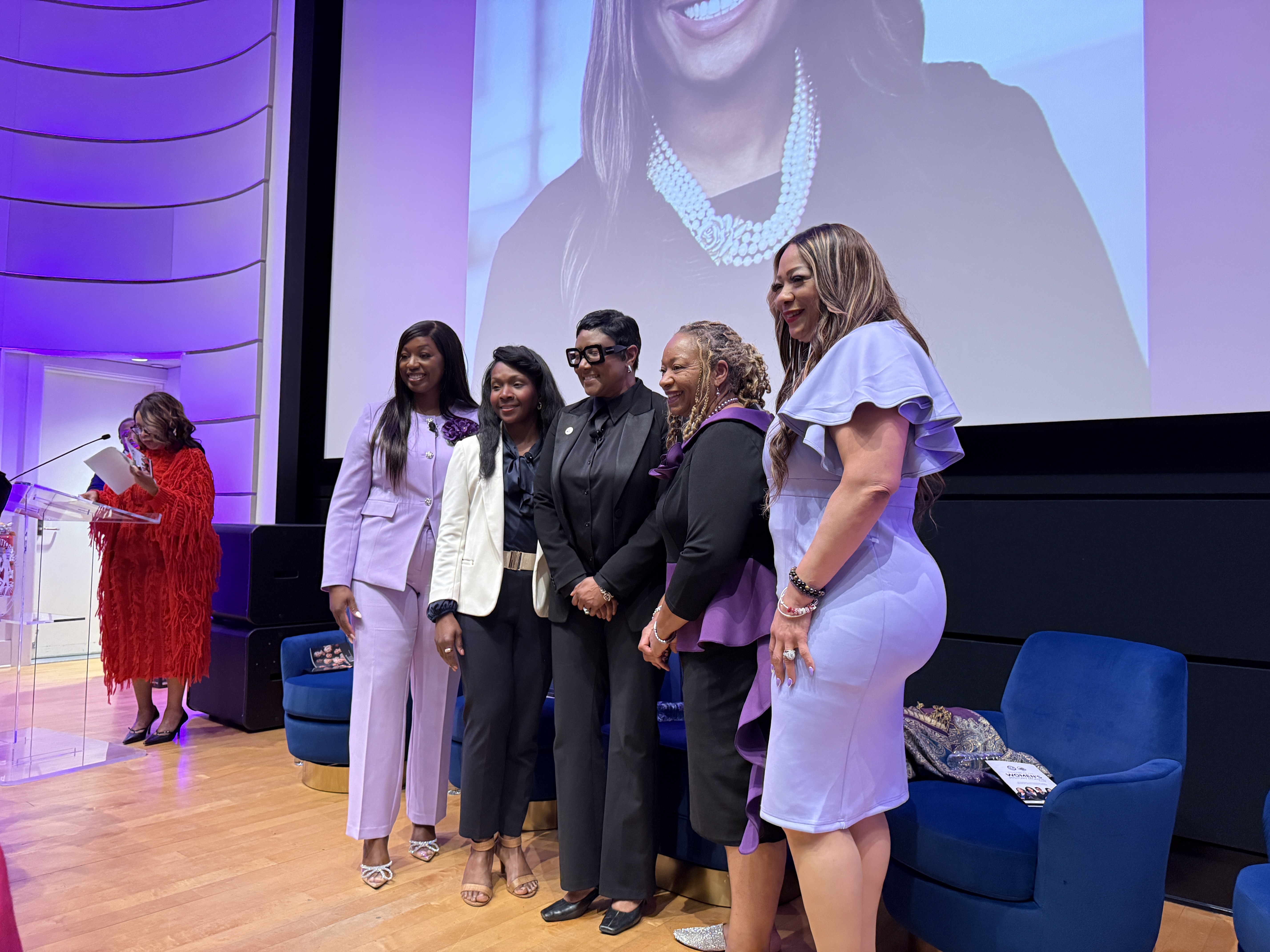 Group of women pose together on stage during a Women’s History Month event. They are dressed in formal attire, including suits and dresses in shades of black, white, and lavender, and stand smiling for a photo. A woman in a red outfit stands at a podium to the left. Behind them, a large screen displays a portrait, and chairs are arranged on stage in a modern auditorium setting.
