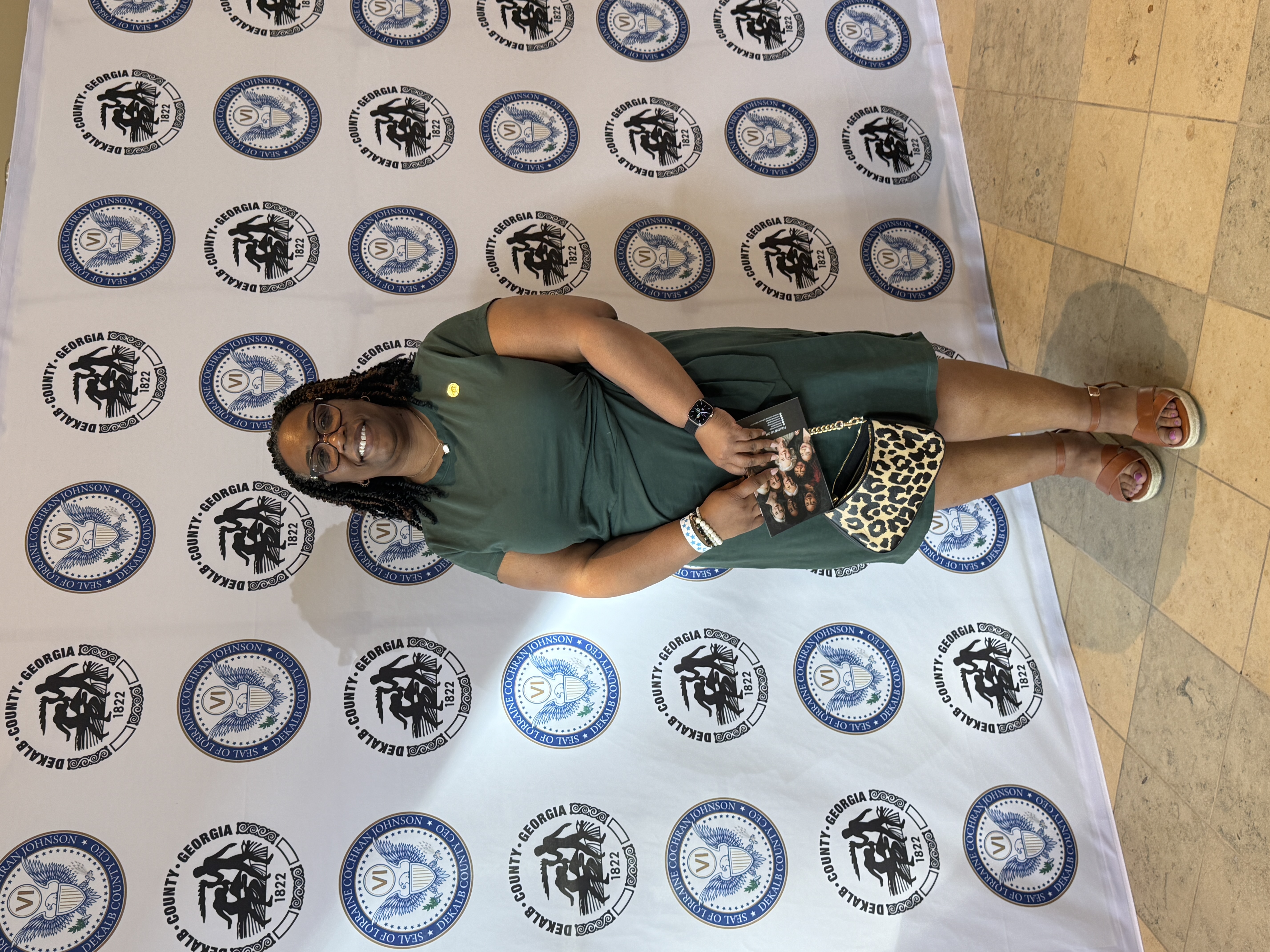 Presiding Officer Commissioner Chakira Johnson stands smiling in front of a step-and-repeat backdrop featuring DeKalb County and CEO Lorraine Cochran-Johnson seals during a Women’s History Month event. She wears a green dress and glasses and holds a small booklet and a leopard-print purse. The setting is indoors with tiled flooring.