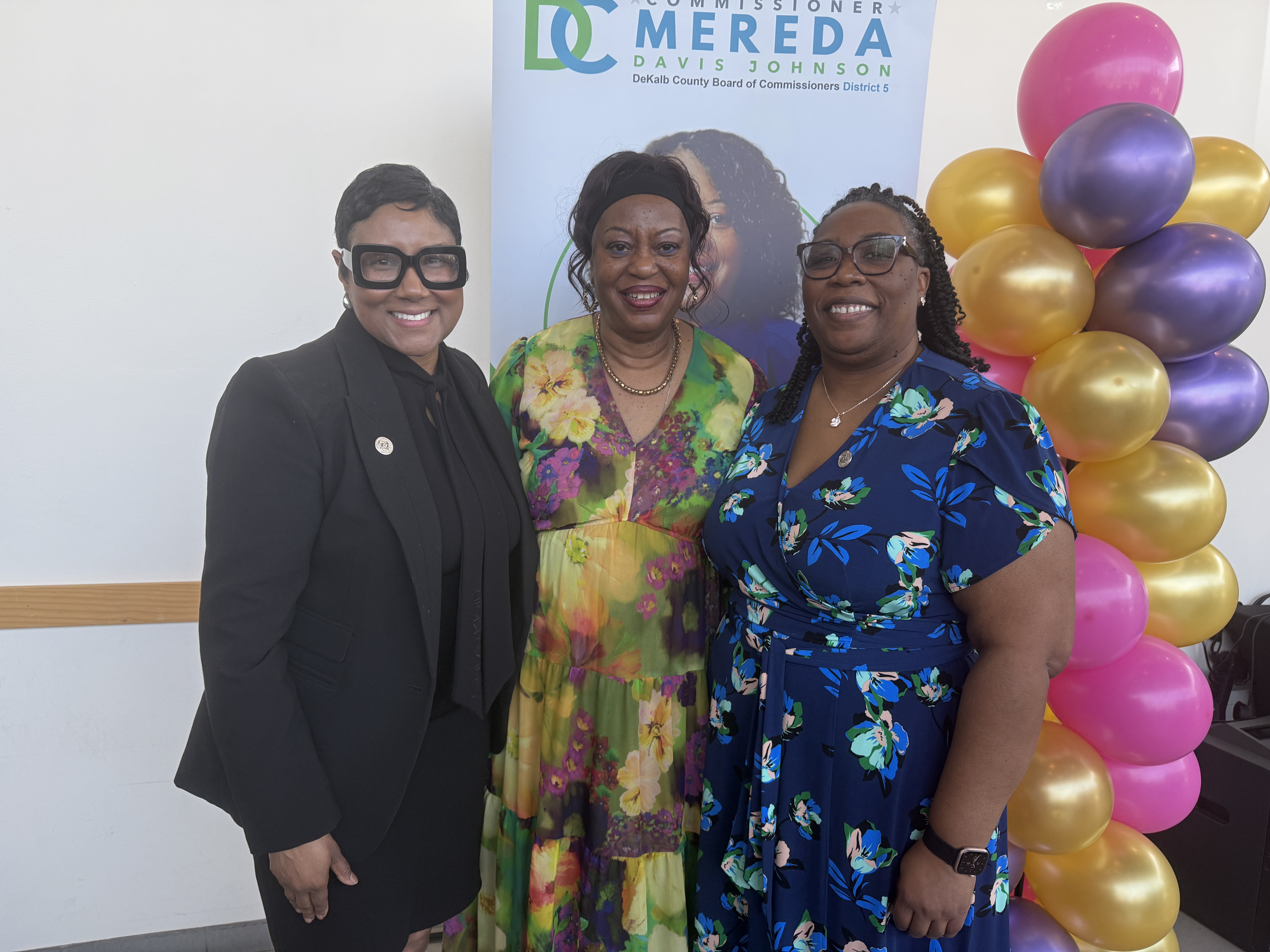 DeKalb County CEO Lorraine Cochran-Johnson, Commissioner Mereda Davis Johnson (District 5), and Commissioner Chakira Johnson (District 4) stand together smiling at an indoor event. CEO Cochran-Johnson wears a black suit and large black glasses. Commissioner Mereda Davis Johnson wears a green and yellow floral dress with a headband. Commissioner Chakira Johnson wears a blue floral dress and glasses. Behind them is a District 5 banner and a column of pink, gold, and purple balloons.
