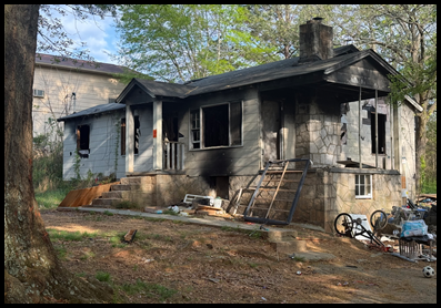 A fire-damaged, abandoned house with boarded and broken windows sits on a lot in Stone Mountain prior to demolition. The structure shows visible burn damage and deterioration, with debris scattered around the yard. The home was located at 5022 West Mountain Street and was demolished on Friday, April 3 at 10:00 a.m. as part of a blight removal effort.