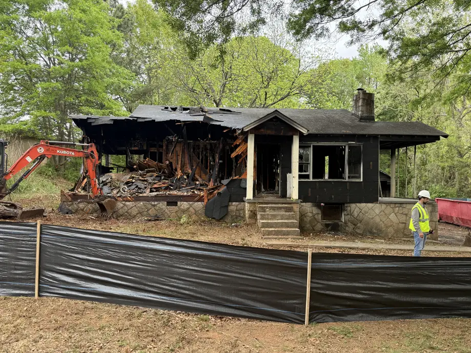 Partially demolished, fire-damaged house at 5022 West Mountain Street in Stone Mountain during a blight removal operation. An excavator tears into the burned structure while a worker in a safety vest and hard hat stands nearby. Debris is visible as the home is actively being taken down. The demolition took place Friday, April 3 at 10:00 a.m.