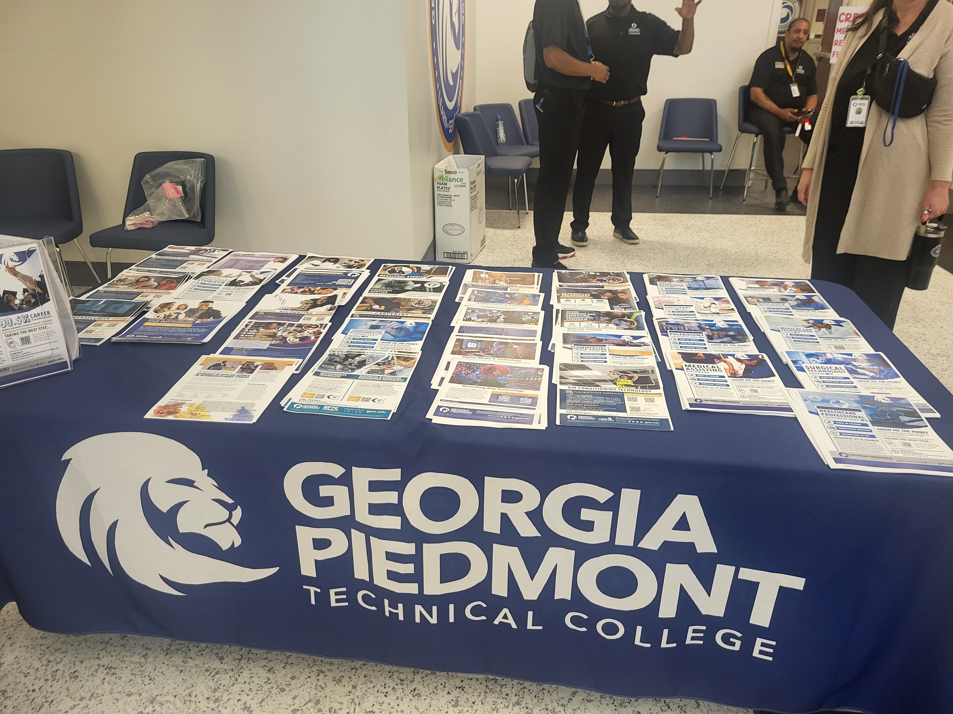 Table display for Georgia Piedmont Technical College at the “Hustle to Higher Education” event, featuring brochures and informational materials about academic programs and career pathways. The table is covered with a branded tablecloth displaying the college’s name and lion logo. Staff and attendees are visible in the background inside a meeting room.