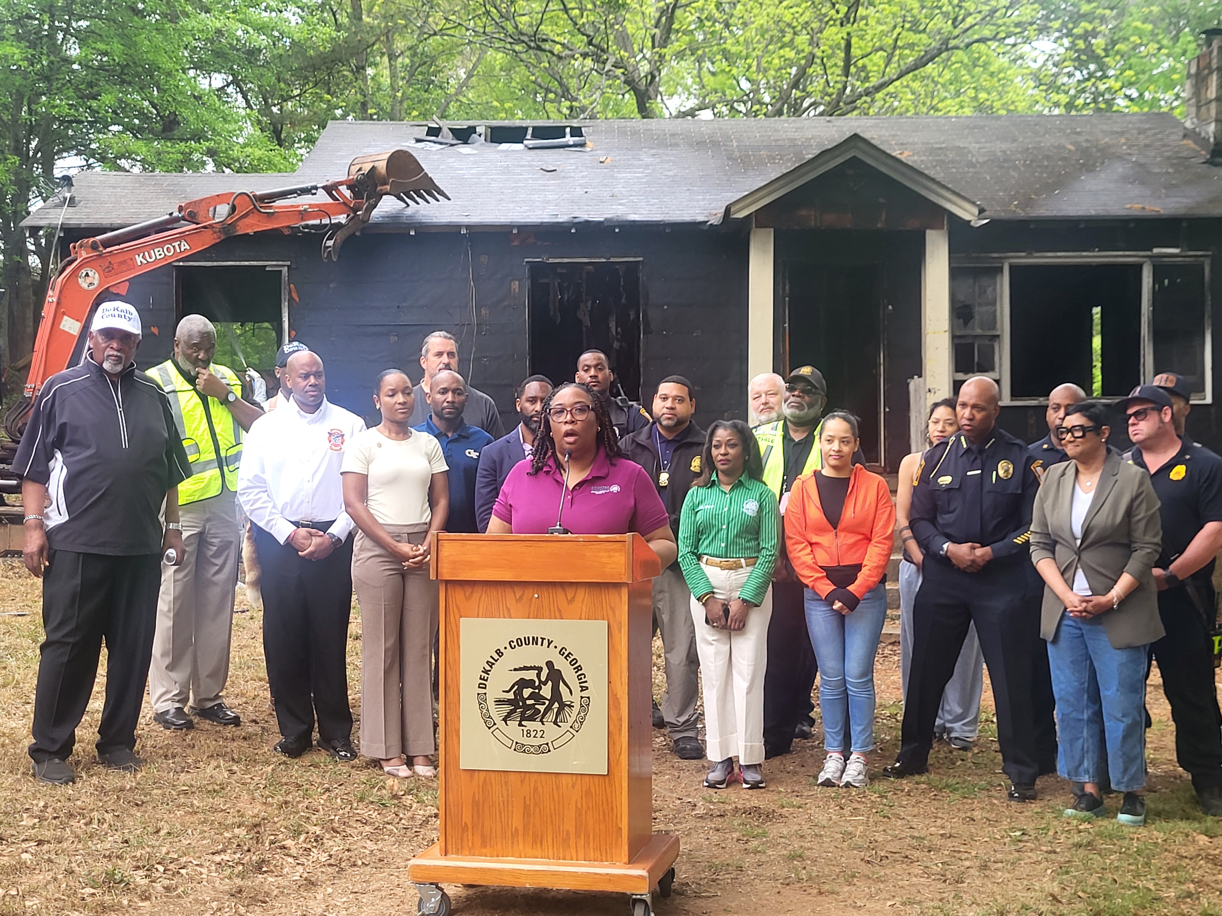 Commissioner Chakira Johnson speaks at a podium during a press event announcing a blight removal project at 5022 West Mountain Street in Stone Mountain. Behind her, DeKalb County officials, public safety personnel, and community partners stand together, including Director of Community Development Allen Mitchell at far left wearing a white baseball cap. A fire-damaged house and an excavator are visible in the background, indicating the ongoing demolition, with additional staff from Community Development present among the group.