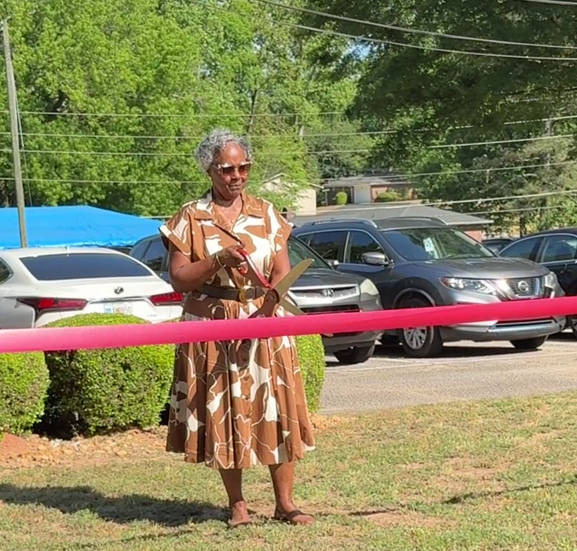 Woman prepares to cut a red ribbon during the Arthur J. Queen Drive renaming ceremony, standing outdoors with vehicles and greenery behind her.