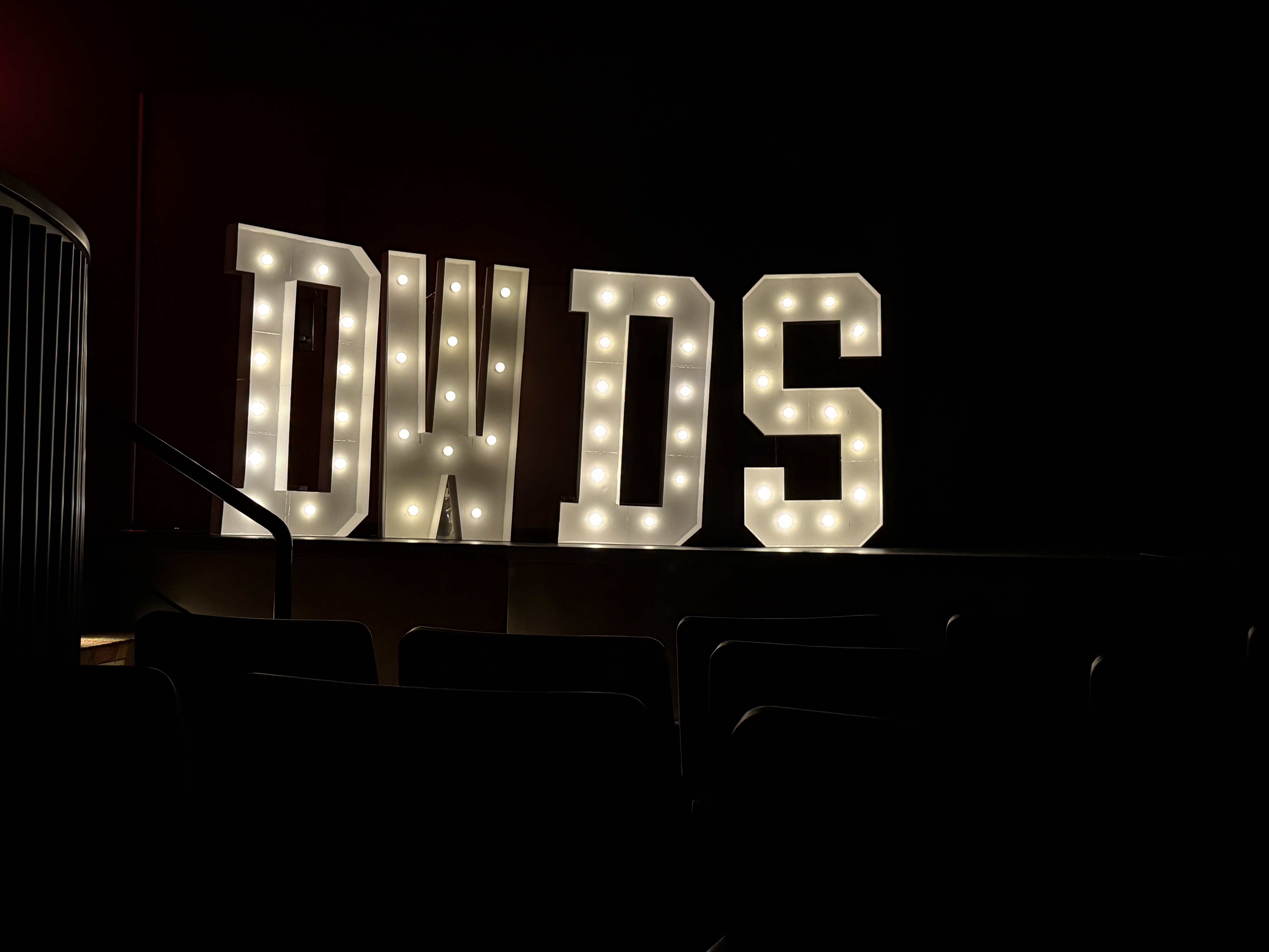 Light-up marquee letters spelling “DWDS” displayed on a stage in a dimly lit auditorium, representing Dancing with the DeKalb Stars.