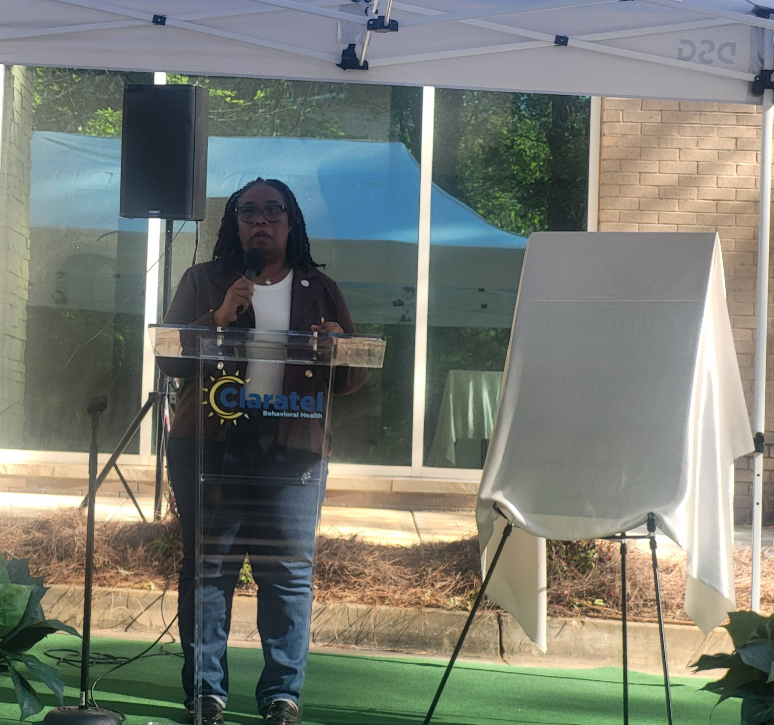 Commissioner Chakira Johnson speaks at a podium during an outdoor event at Claratel Behavioral Health, standing beside a covered display prepared for unveiling.