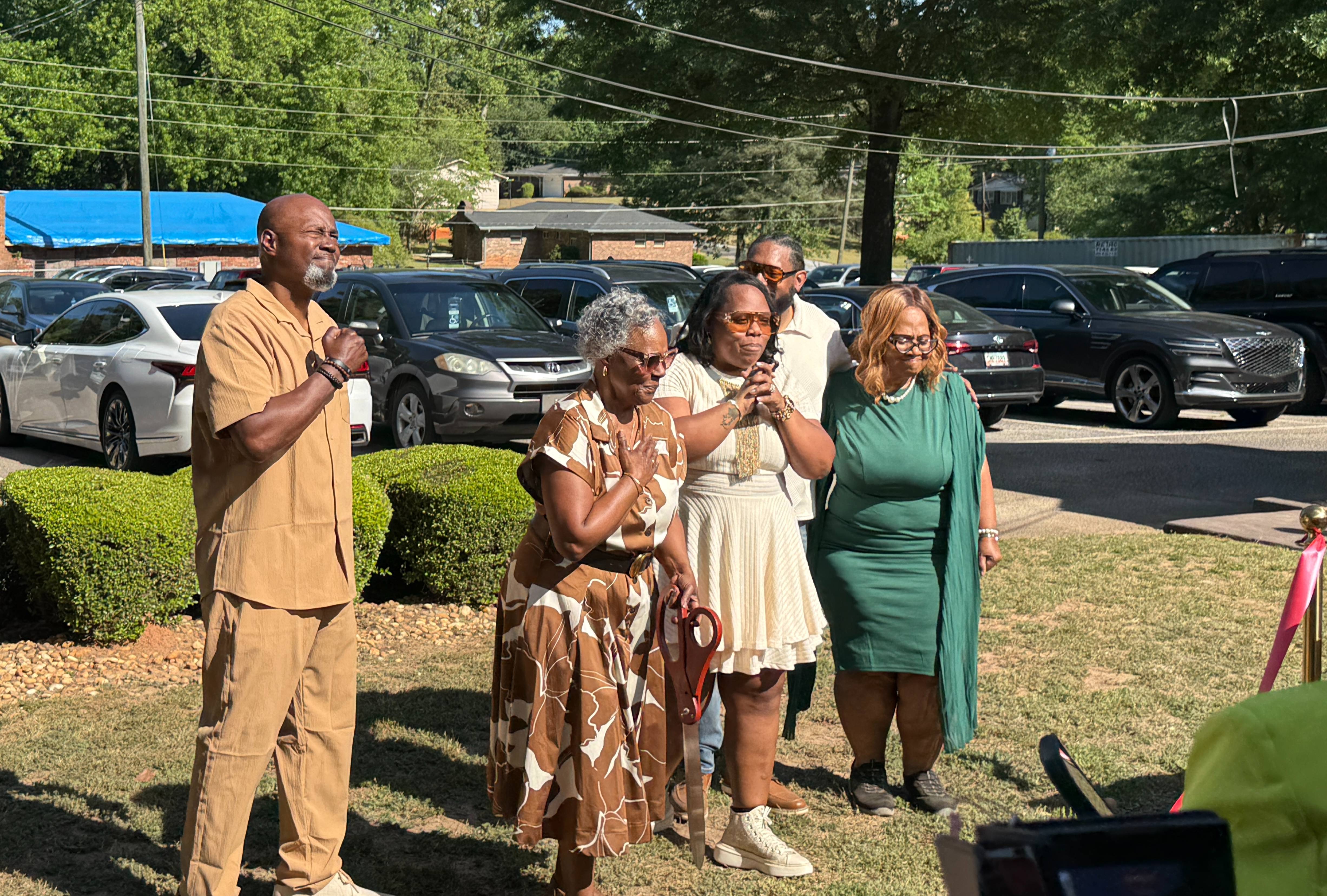 Members of Arthur J. Queen’s family stand together during an outdoor ribbon-cutting ceremony, some with hands over their hearts and others clasping hands, as one person holds ceremonial scissors.