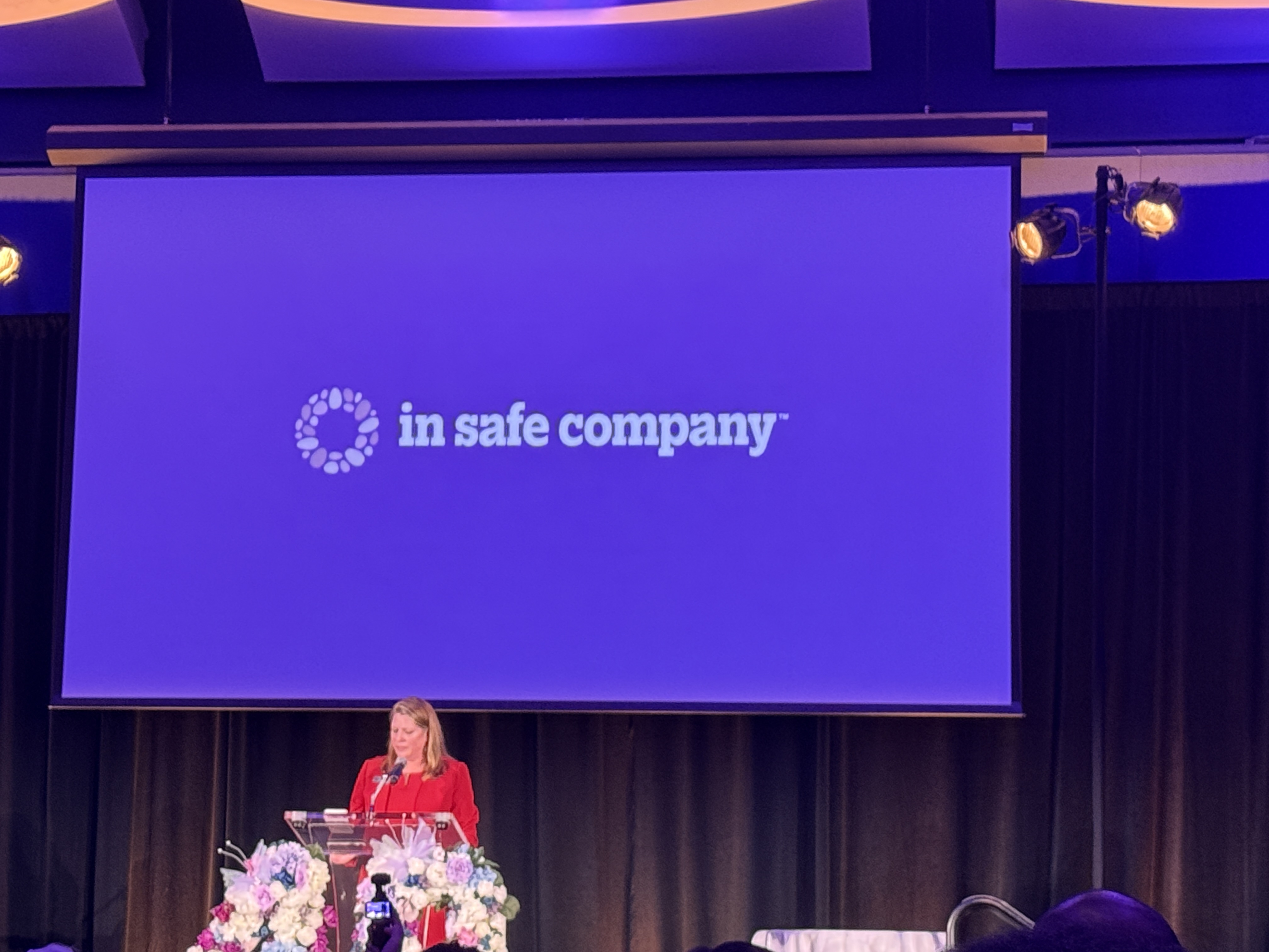 A speaker stands at a podium onstage during the Women’s Resource Center to End Domestic Violence’s Champions for Change gala. Above the stage, a large projection screen displays the organization’s new “in safe company” branding as part of its rebrand announcement. Purple lighting and floral arrangements frame the stage.