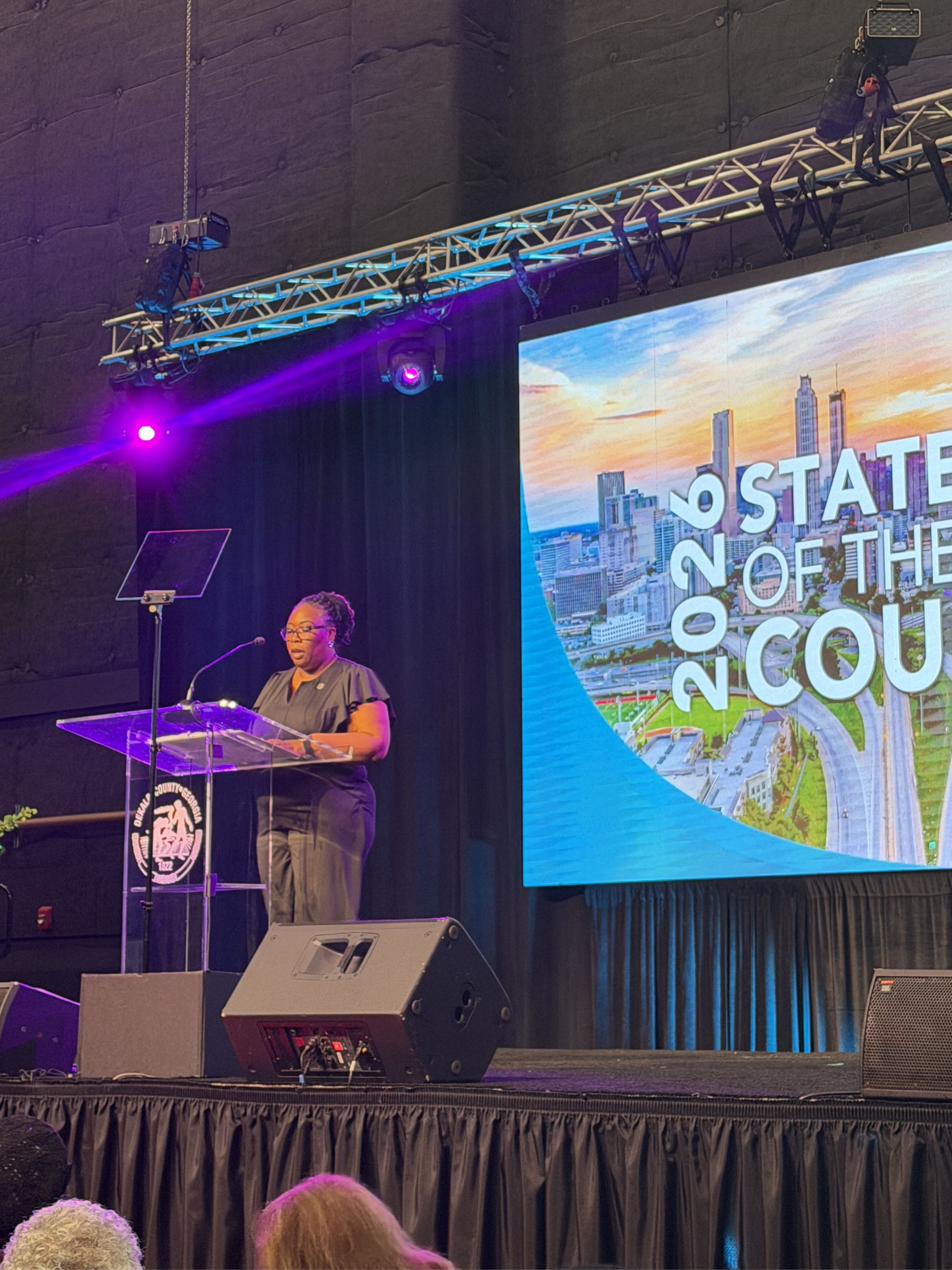Presiding Officer District 4 Commissioner Chakira Johnson stands at a podium delivering remarks during DeKalb County’s 2026 State of the County Address. A large digital screen displaying “2026 State of the County” and an Atlanta skyline graphic is visible beside the stage. Purple and blue stage lighting illuminate the venue as attendees watch from the audience.