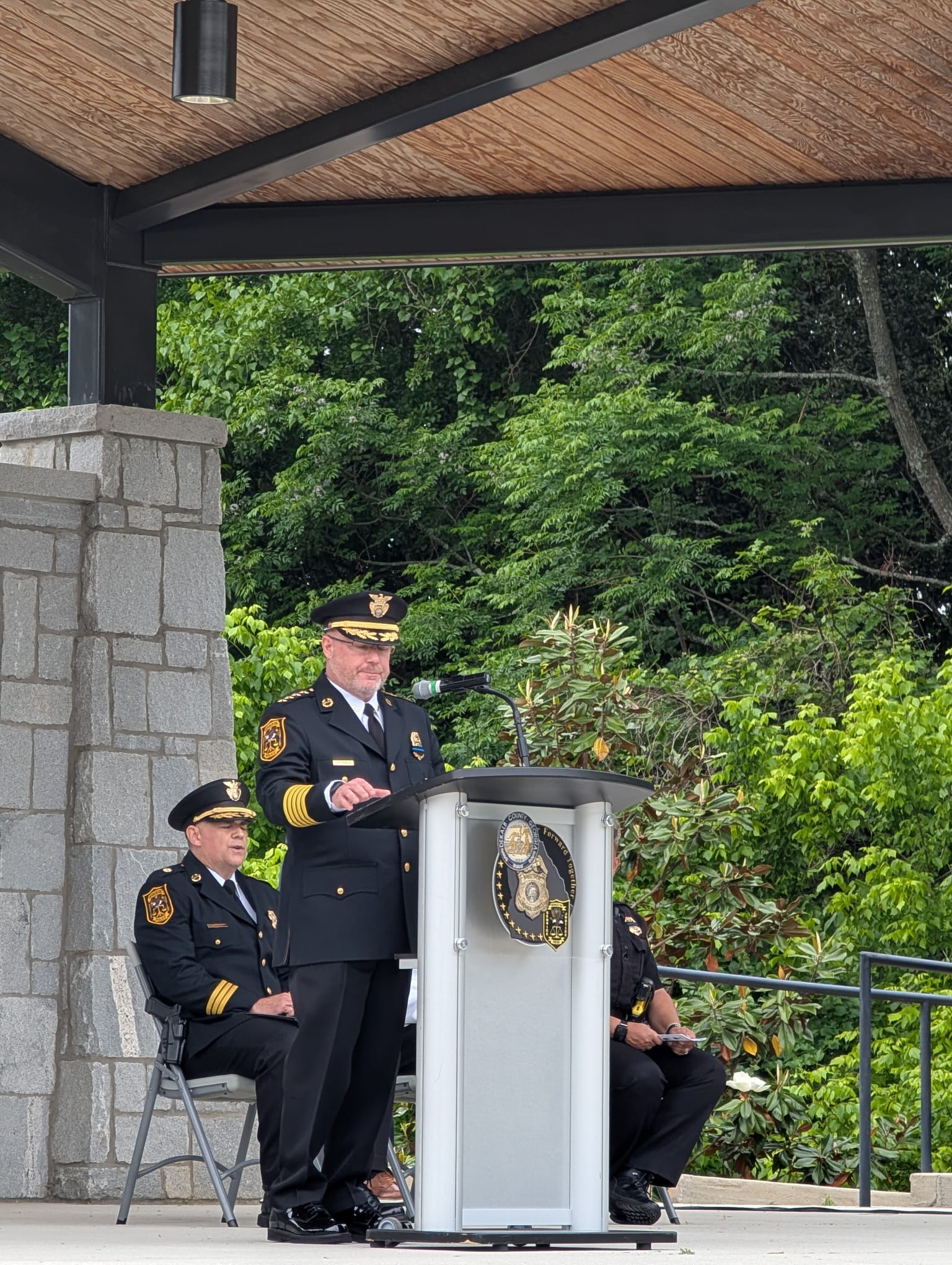 DeKalb Police Chief Gregory Padrick stands at a podium delivering remarks during the Fallen Officers Memorial Ceremony. Uniformed officers are seated behind him beneath an outdoor pavilion surrounded by trees and greenery. The podium displays DeKalb County Police Department insignia as attendees gather to honor fallen officers and recognize their service and sacrifice.