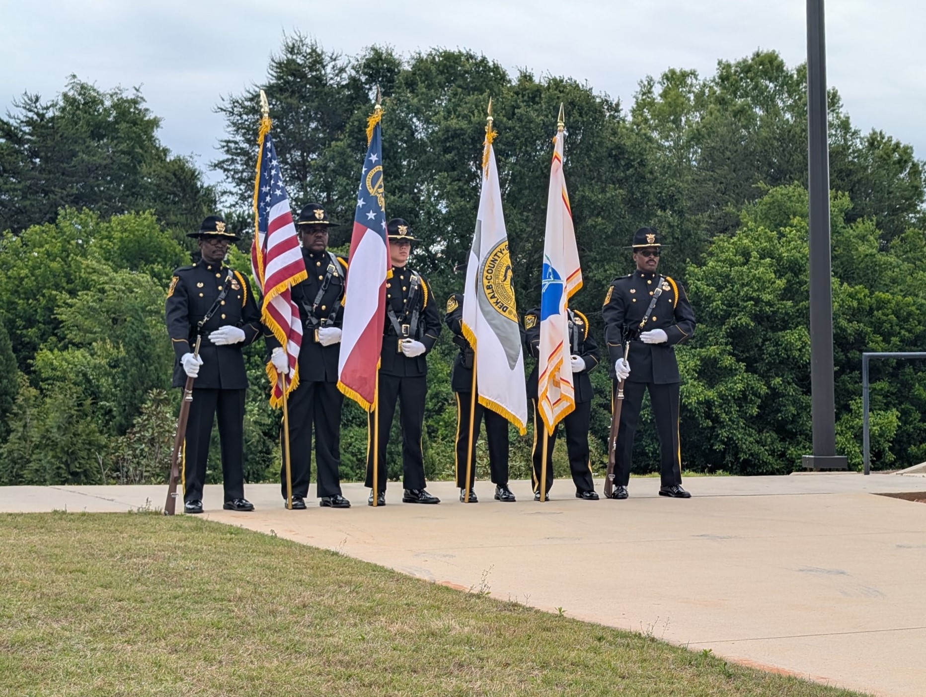 Uniformed members of a ceremonial honor guard stand in formation during the Fallen Officers Memorial Ceremony. Officers hold the American flag, the Georgia state flag, and additional ceremonial flags while standing outdoors near a pavilion surrounded by trees and greenery.
