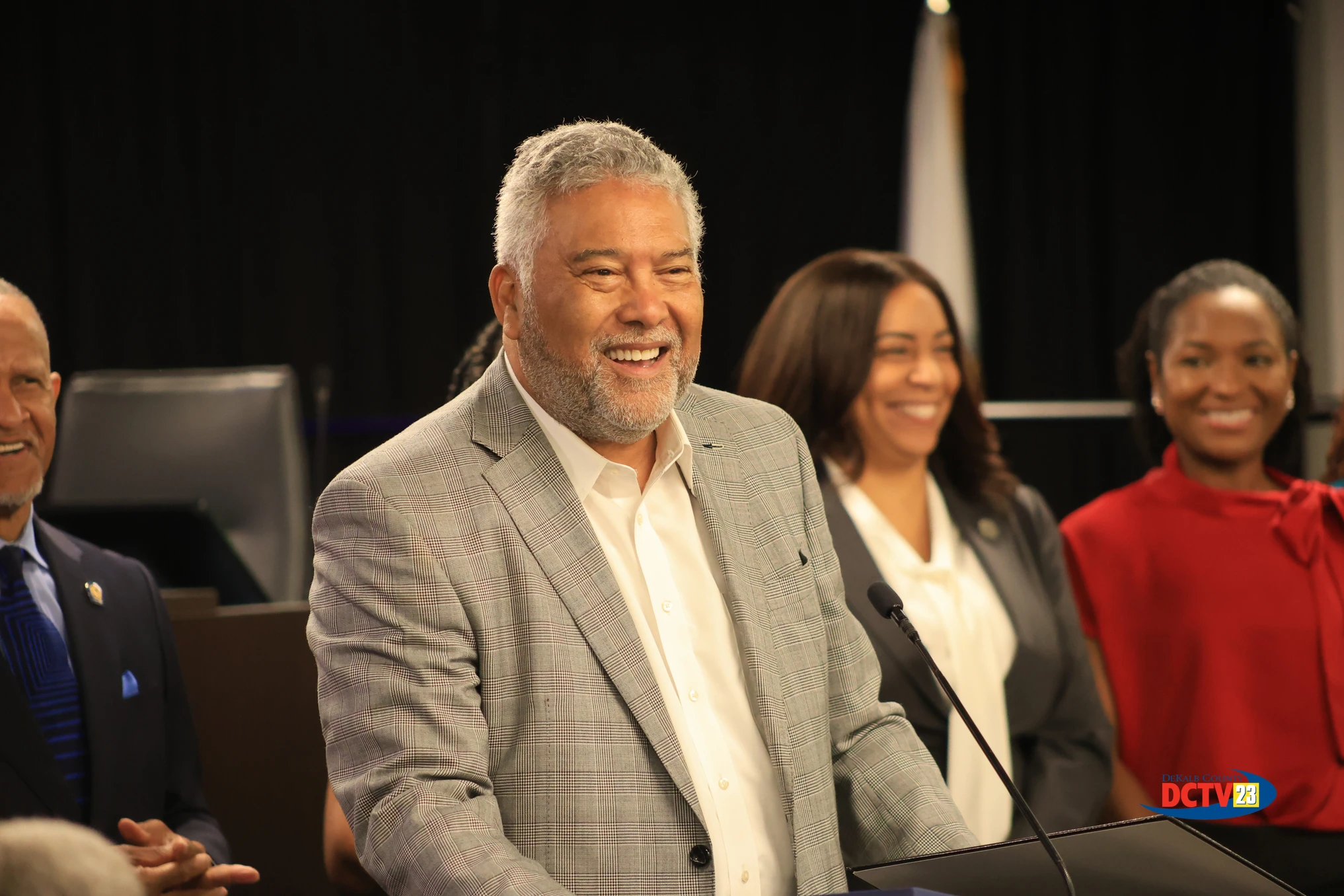 Former DeKalb Sheriff and retired U.S. Marshal Thomas E. Brown smiles while speaking at a podium during a special recognition presentation at a DeKalb County Board of Commissioners meeting. Other attendees and county officials stand behind him inside the meeting chamber. Photo: DCTV23.