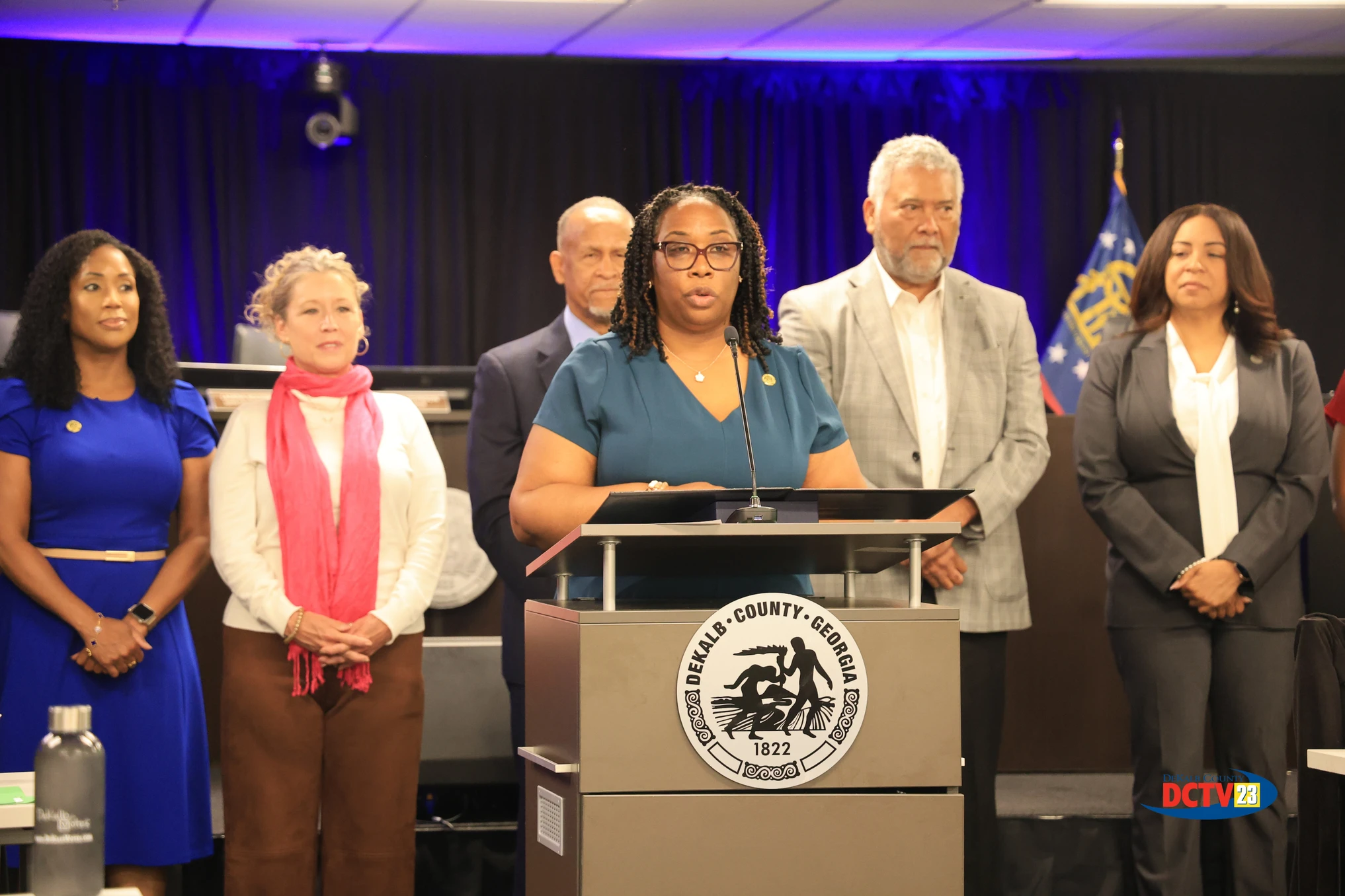 Presiding Officer Commissioner Chakira Johnson speaks at a podium during a special recognition held at a DeKalb County Board of Commissioners regular meeting honoring longtime public servants and community leaders. Standing behind her from left to right are District 3 Commissioner Nicole Massiah, District 2 Commissioner Michelle Long Spears, former DeKalb Tax Commissioner Irvin Johnson, former DeKalb Sheriff and retired U.S. Marshal Thomas E. Brown, and newly appointed DeKalb Tax Commissioner Nicole Golden. The meeting takes place inside the county chamber with blue accent lighting and a DeKalb County seal displayed on the podium. Photo: DCTV23.