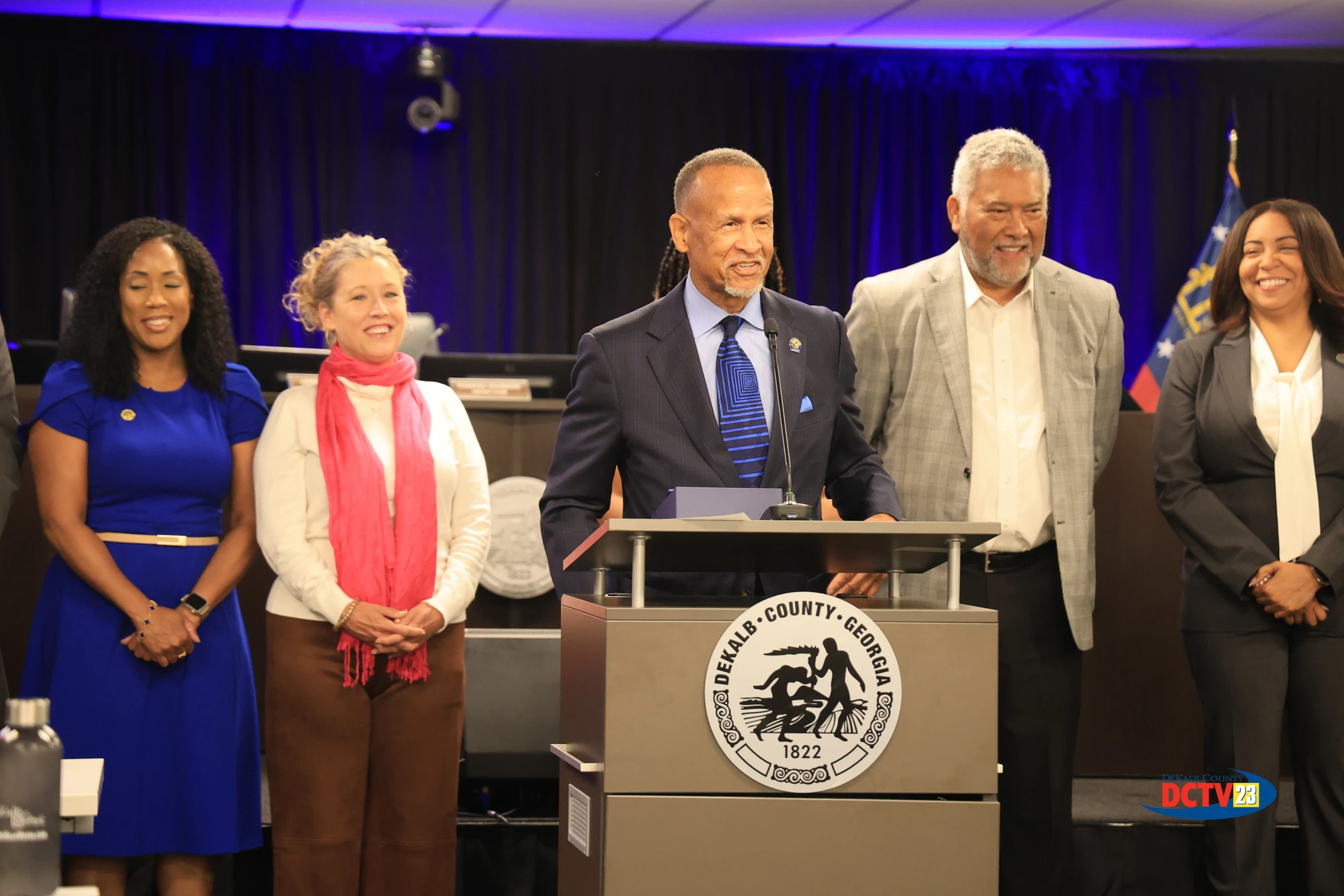 Former DeKalb Tax Commissioner Irvin Johnson speaks at a podium during a special recognition presentation at a DeKalb County Board of Commissioners meeting while attendees and county officials stand behind him. DeKalb's current Tax Commissioner Nicole Golden and former U.S. Marshal Thomas E. Brown are visible among the group. Photo: DCTV23.