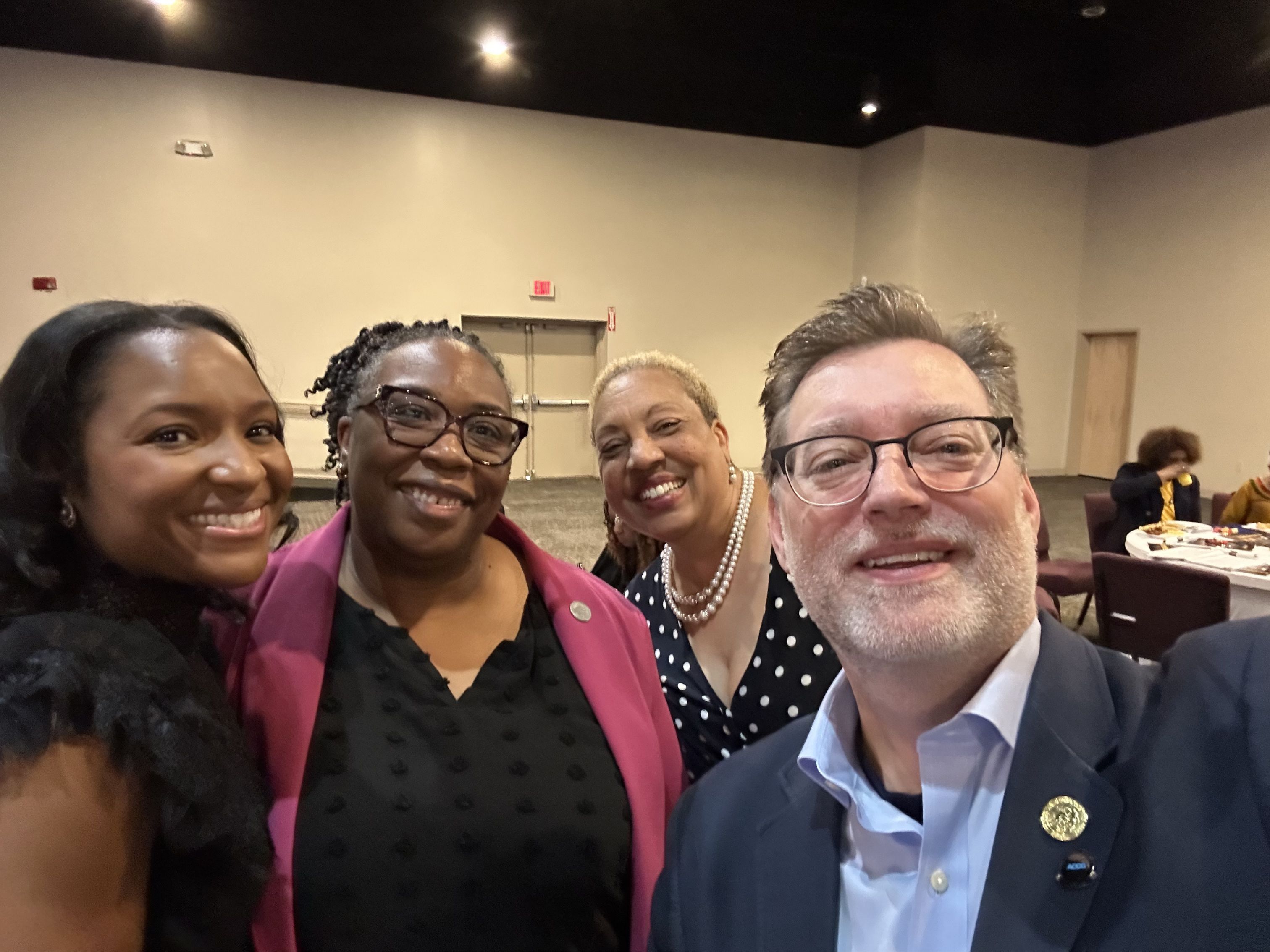District 1 Commissioner Robert Patrick takes a selfie with Deputy Presiding Officer Super District 7 Commissioner Dr. LaDena Bolton, Presiding Officer Commissioner Chakira Johnson, and another attendee during the NAACP DeKalb County Branch Unity Breakfast. The group smiles inside the event hall with banquet tables visible in the background.