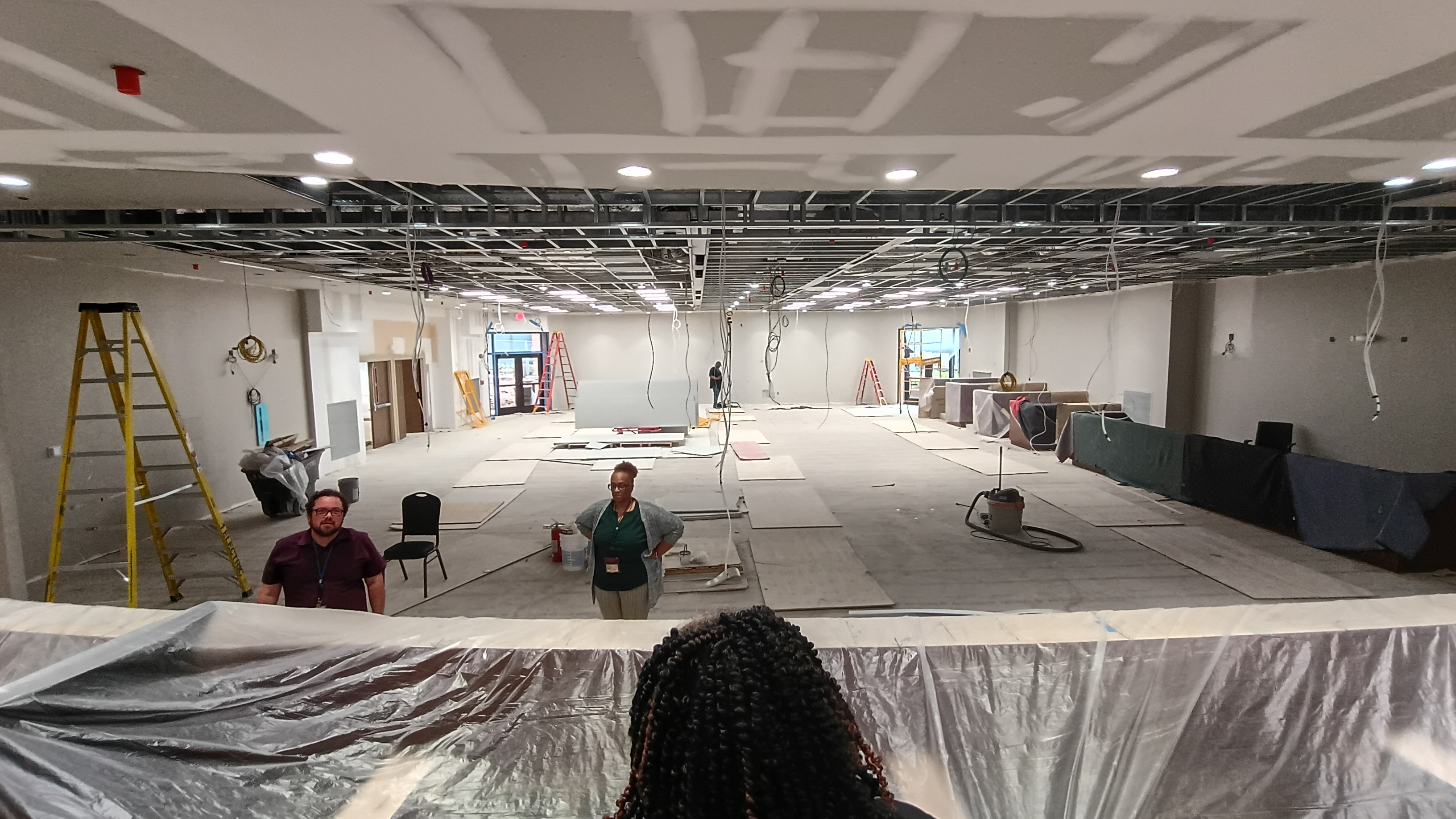 Wide view of the Maloof Center auditorium during renovation construction, photographed from the dais area. The large room has exposed ceiling framework, hanging electrical wires, ladders, construction materials, and partially installed drywall and flooring. Several people stand inside the unfinished space while work continues throughout the auditorium.