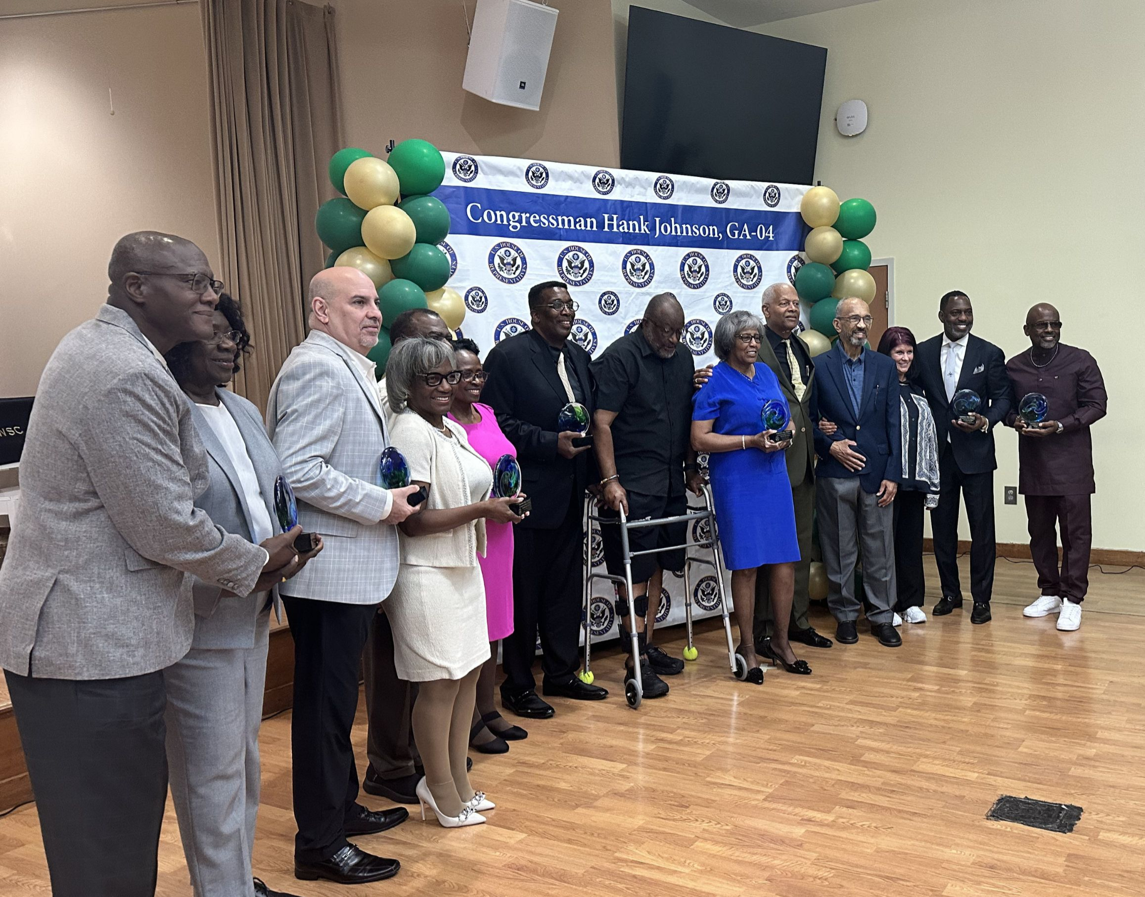 Group photo from Congressman Hank Johnson’s Pinnacle Awards event. A line of award recipients and attendees stand together in front of a “Congressman Hank Johnson, GA-04” backdrop decorated with green and gold balloons. Several people hold glass award trophies while posing for the photo inside an event hall.
