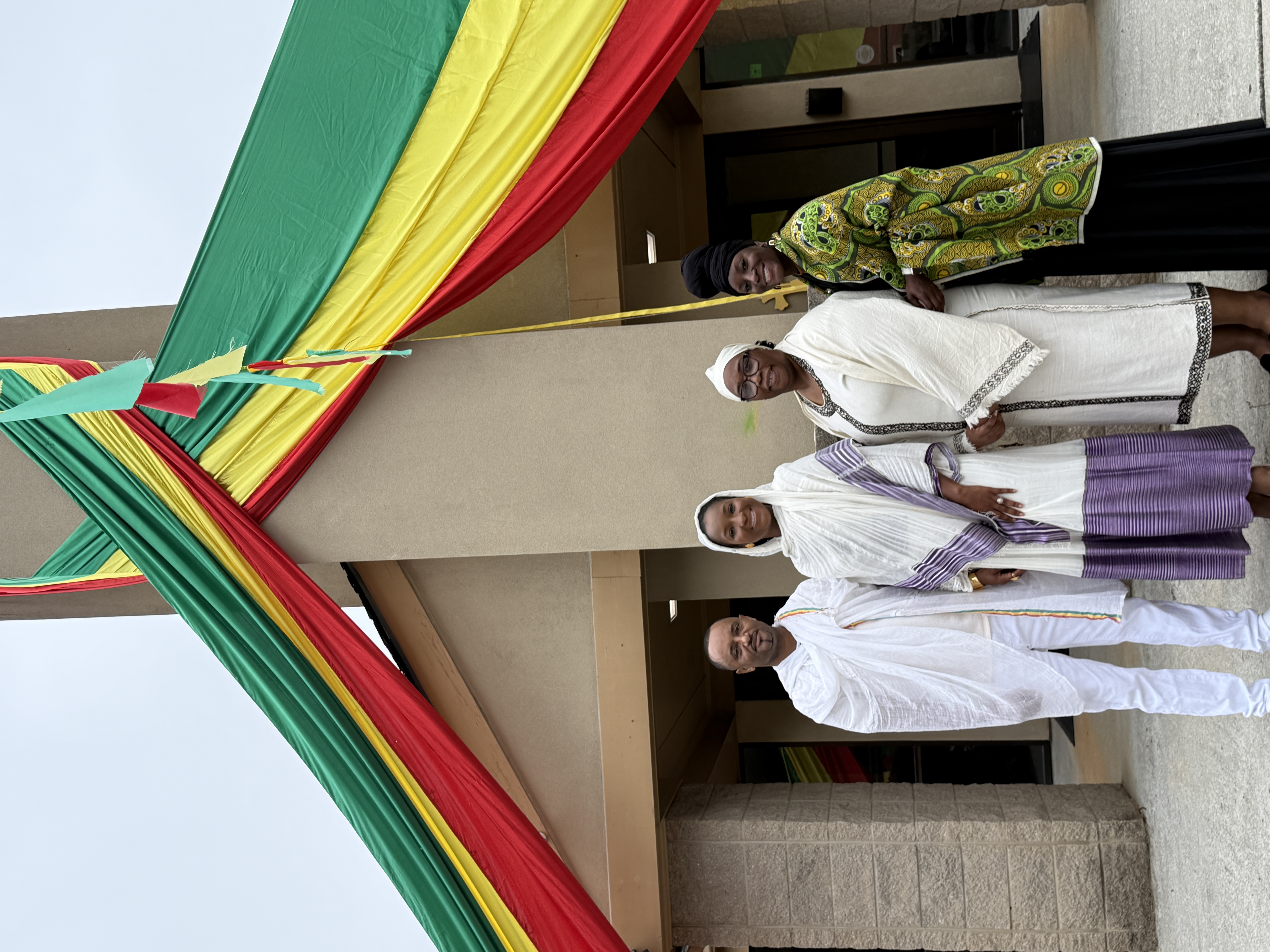 Four individuals stand outside the Mahidere Sibhat Lideta LeMariam Monastery EOTC beneath red, yellow, and green fabric draped across the building entrance during an annual monastery celebration in May 2026. From left to right are an unidentified man dressed in traditional white attire, Deputy Presiding Officer Super District 7 Commissioner Dr. LaDena Bolton, Presiding Officer Commissioner Chakira Johnson, and an unidentified woman in traditional patterned clothing.