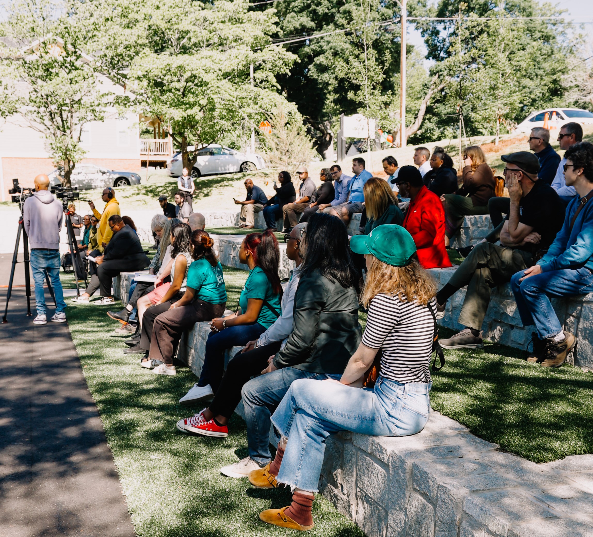 Audience members sit on stone seating during the Cedar Park ribbon cutting event. Attendees watch speakers and camera crews gathered near the front of the park’s amphitheater-style seating area.