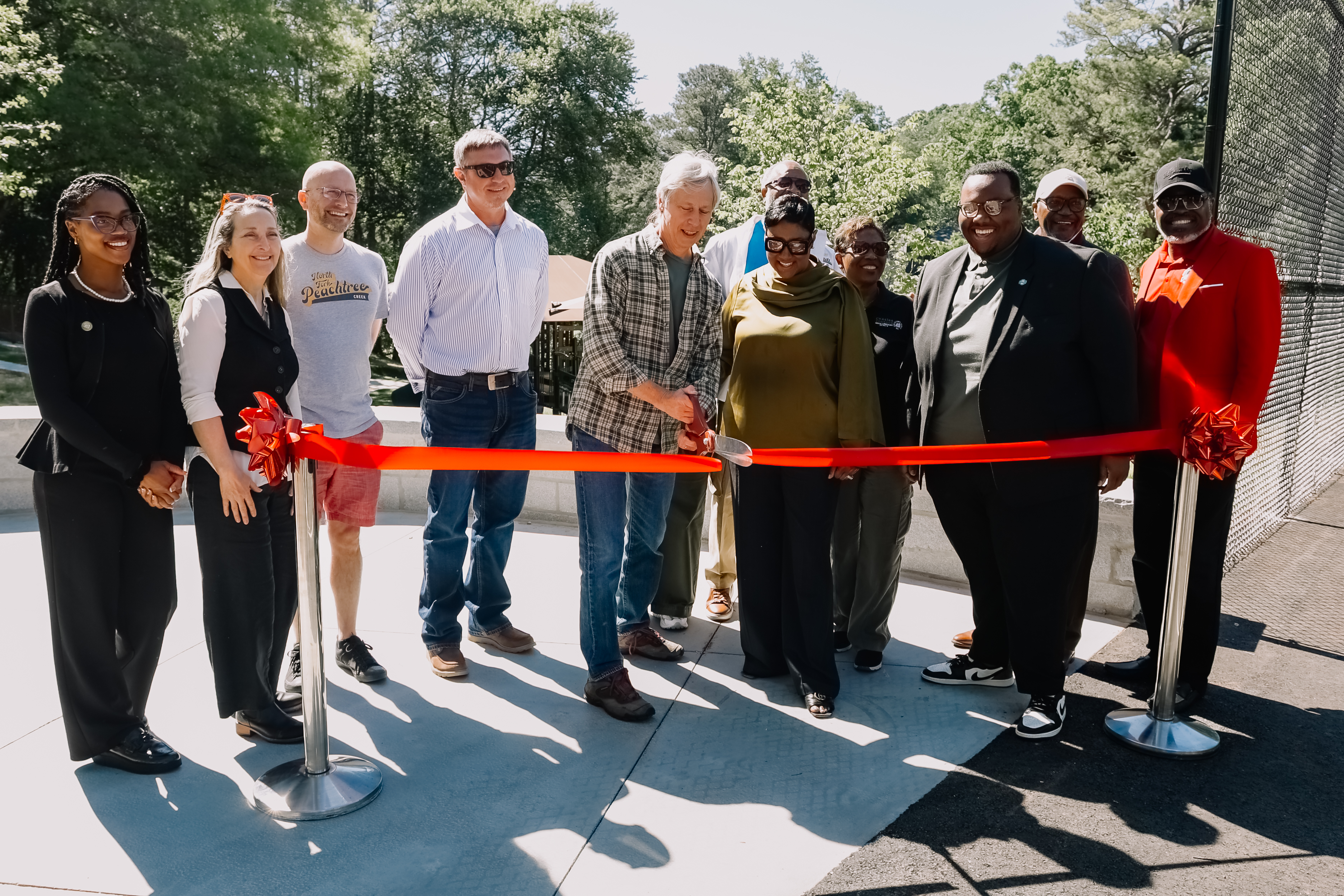 Group photo at a ribbon cutting ceremony at Cedar Park. Several community leaders and attendees stand behind a red ribbon outdoors on a sunny day while one person cuts the ribbon. Trees and park features are visible in the background.