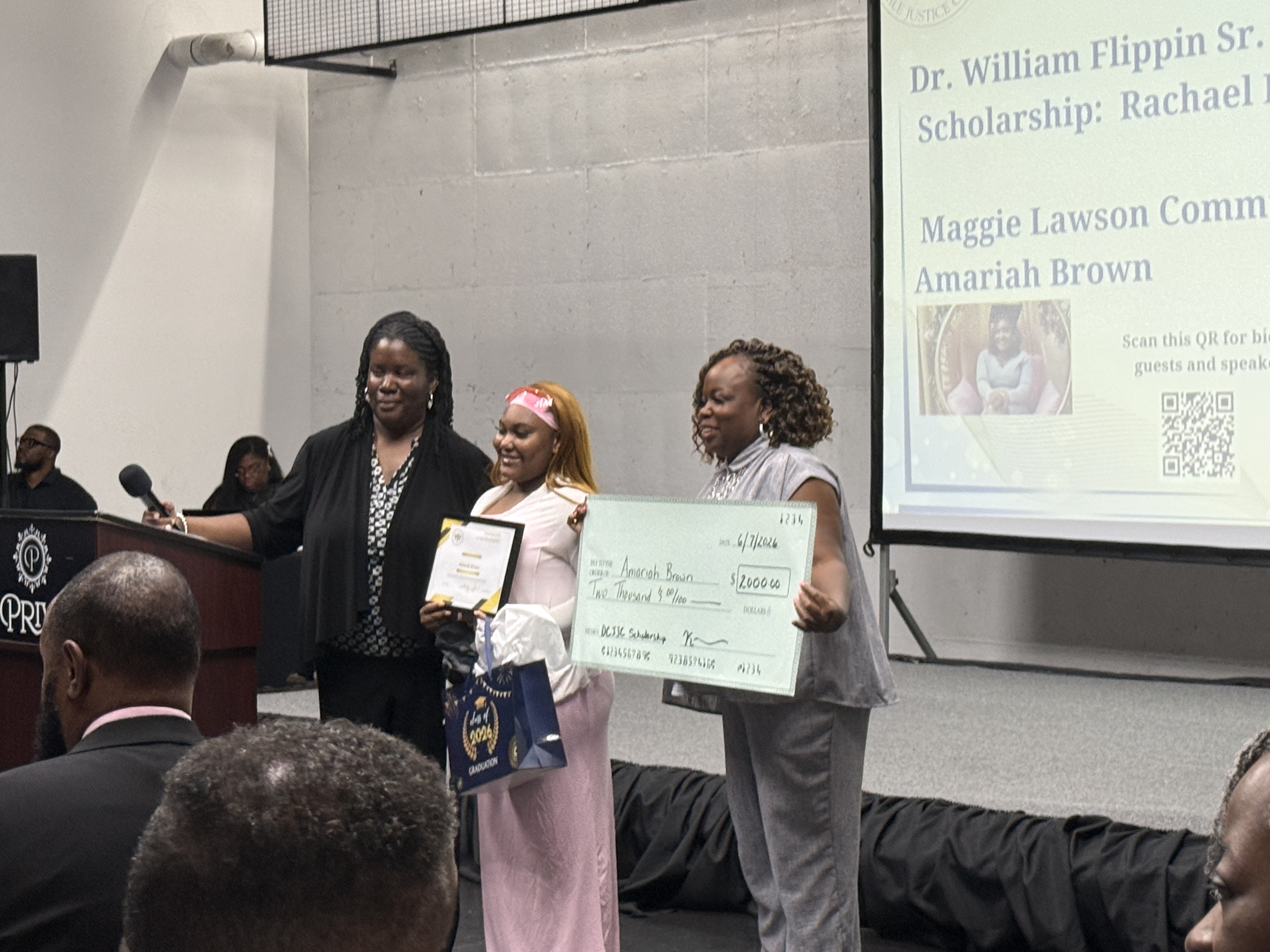 A scholarship recipient stands with community leaders while receiving a certificate and ceremonial check during a DeKalb County Juvenile Justice Council recognition event. A presentation screen is visible in the background.
