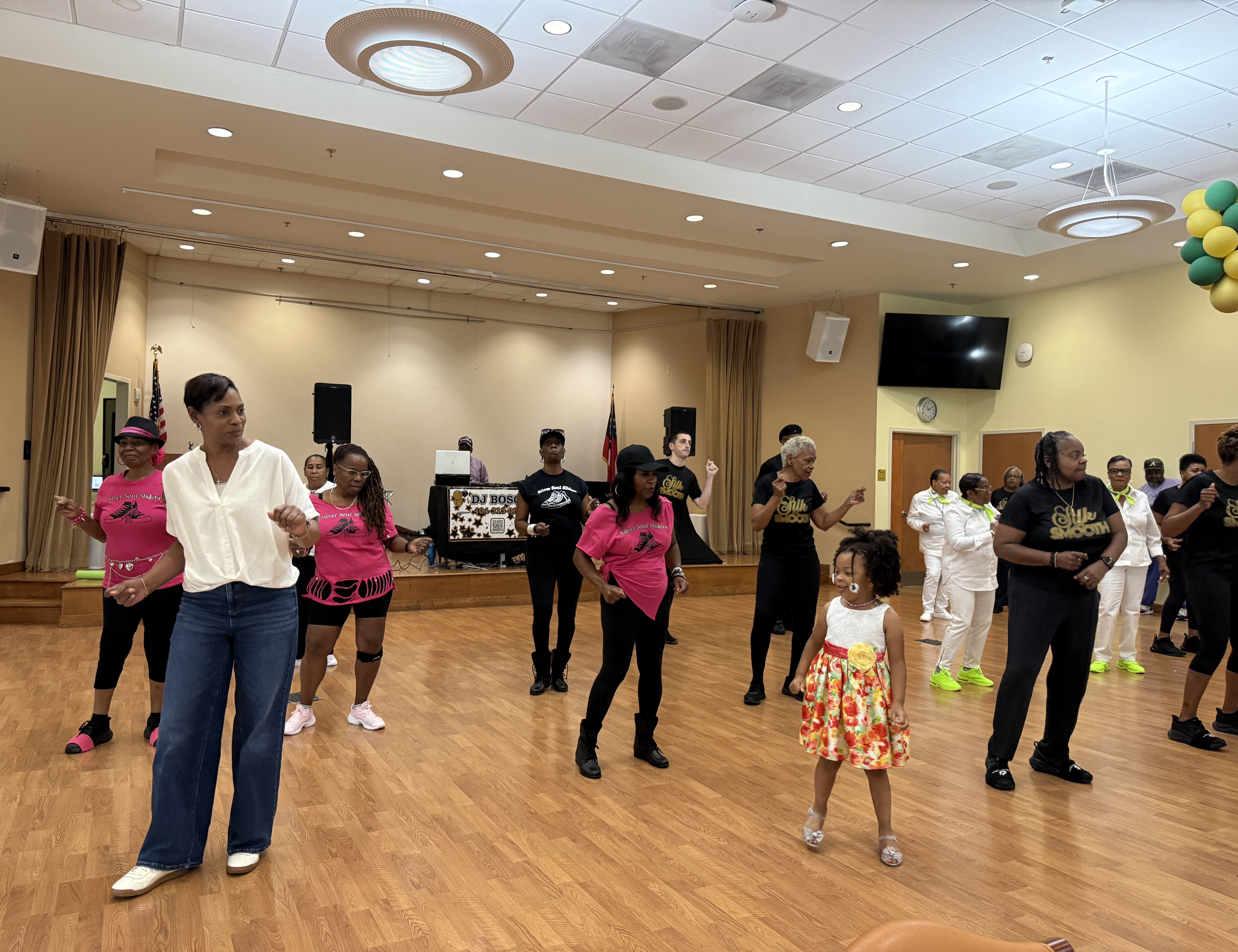 Community members participate in a group dance during the Dancing for Dementia event at the Lou Walker Senior Center. Attendees of various ages gather on the dance floor for a line dance activity while music plays in the background.