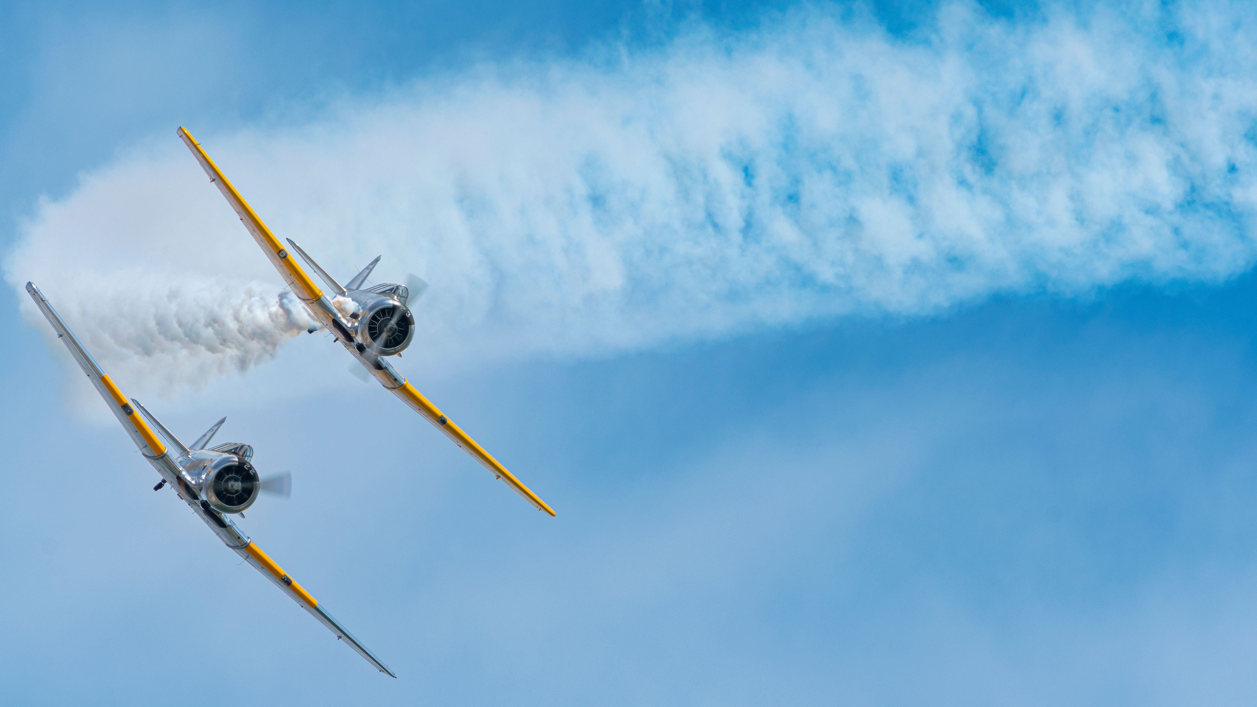 Two vintage propeller aircraft perform an aerial maneuver against a blue sky, with one plane leaving a trail of white smoke.