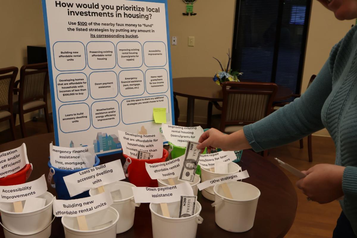 A participant places play money into labeled buckets during an interactive housing priorities activity. The display asks participants to prioritize local housing investments among options such as affordable housing, home repairs, down payment assistance, accessibility improvements, and emergency financial assistance. The image represents a previous community engagement activity related to housing planning.