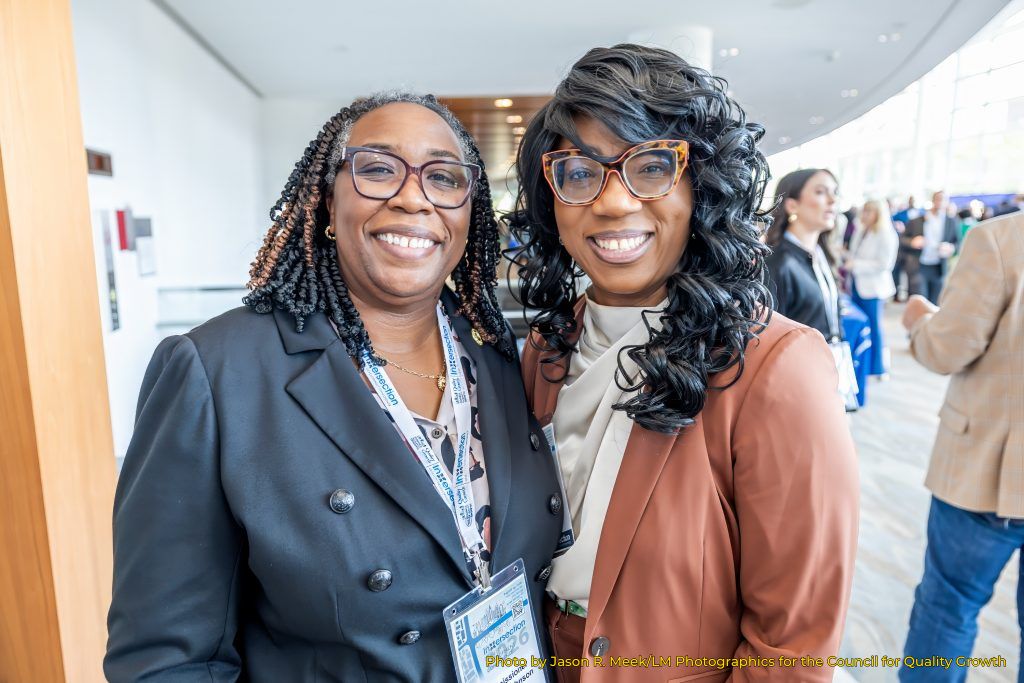 Presiding Officer Commissioner Chakira Johnson smiles alongside another conference attendee in a bright networking area during the INTERSECTION Quality Development Conference, with other attendees gathered in the background.