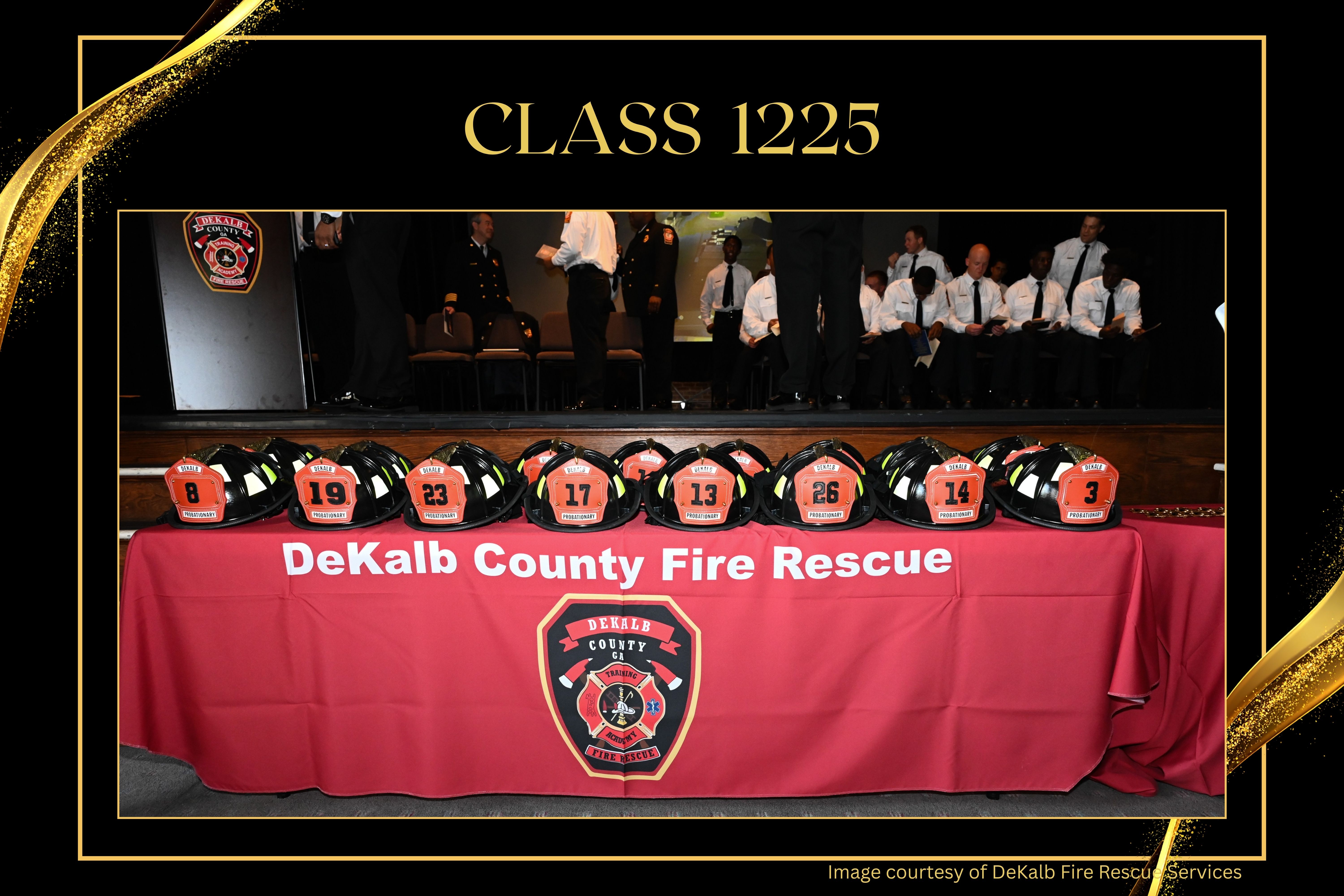 A row of black firefighter helmets with orange DeKalb County Fire Rescue shields sits on a table draped with a red DeKalb County Fire Rescue banner. Members of Class 1225 are seated onstage in the background during the graduation ceremony.