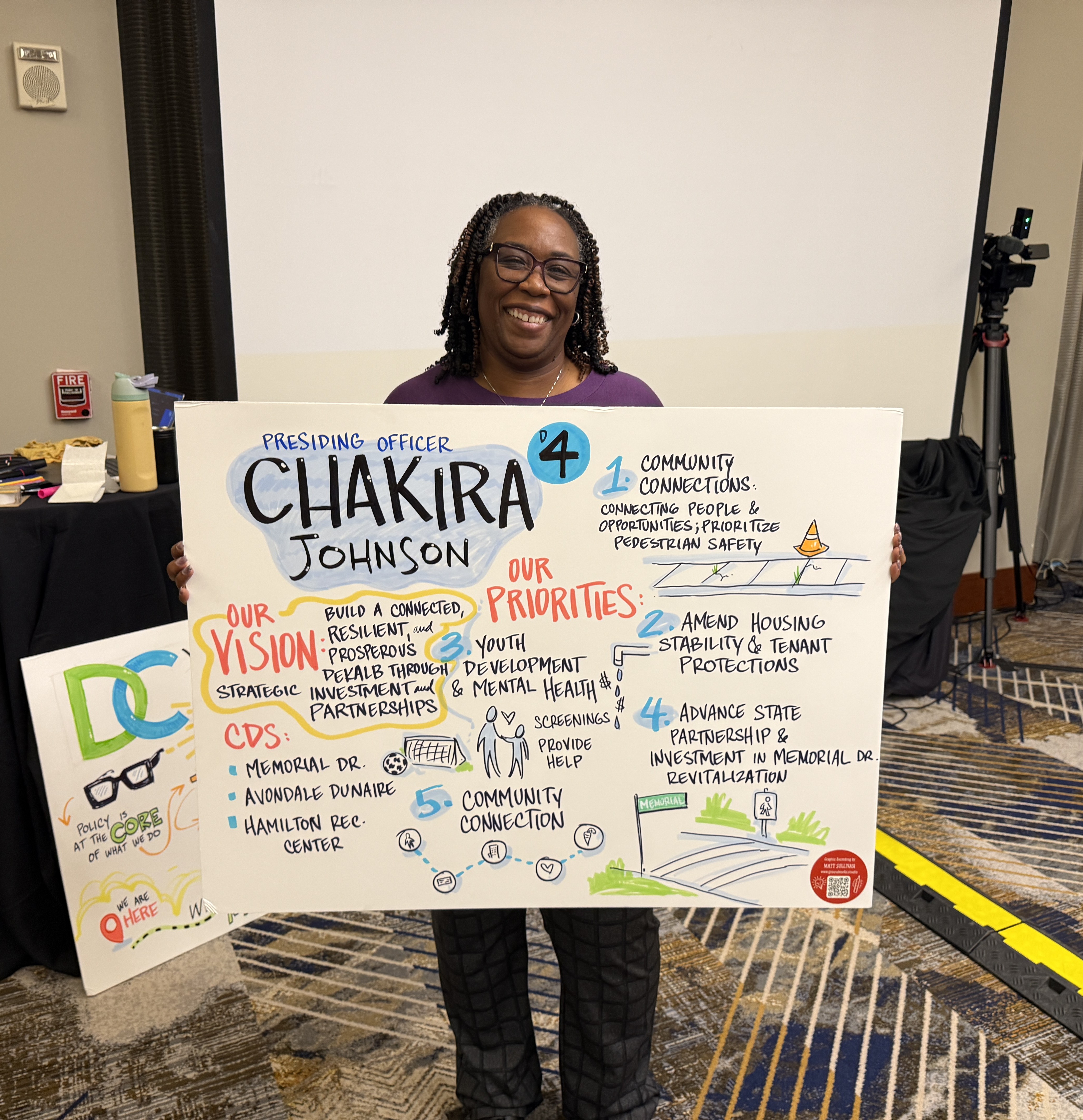 Presiding Officer Commissioner Chakira Johnson smiles while holding a hand-lettered poster outlining District 4’s vision and 2026–2027 priorities. The poster highlights community connections and pedestrian safety, housing stability and tenant protections, youth development and mental health, Memorial Drive revitalization, and community connection, along with congressional funding priorities for Memorial Drive, Avondale Dunaire Park and Hamilton Recreation Center.