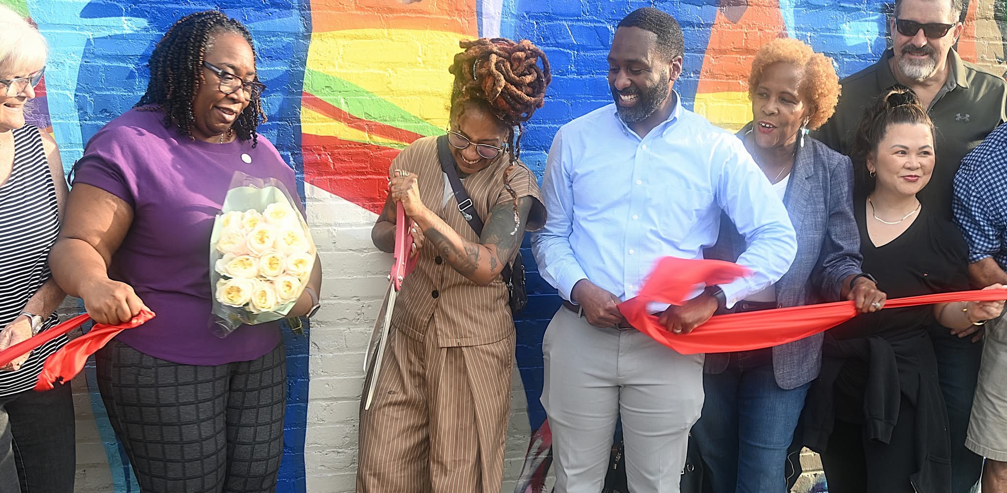 Presiding Officer Commissioner Chakira Johnson stands with a bouquet of white roses as visual artist Charity Hamidullah cuts a red ribbon beside Stone Mountain Mayor Jelani Linder and other community members in front of the new mural.