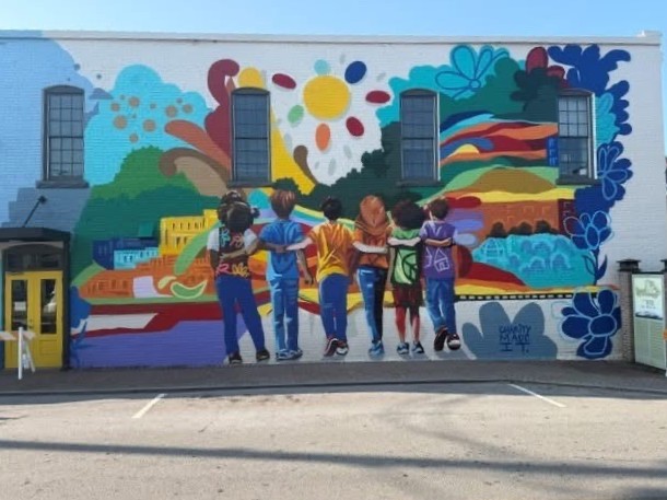 Large colorful mural painted on the side of a downtown Stone Mountain building. The artwork shows six children standing shoulder to shoulder with their backs to the viewer, facing a bright landscape filled with flowers, trees, buildings and a yellow sun.