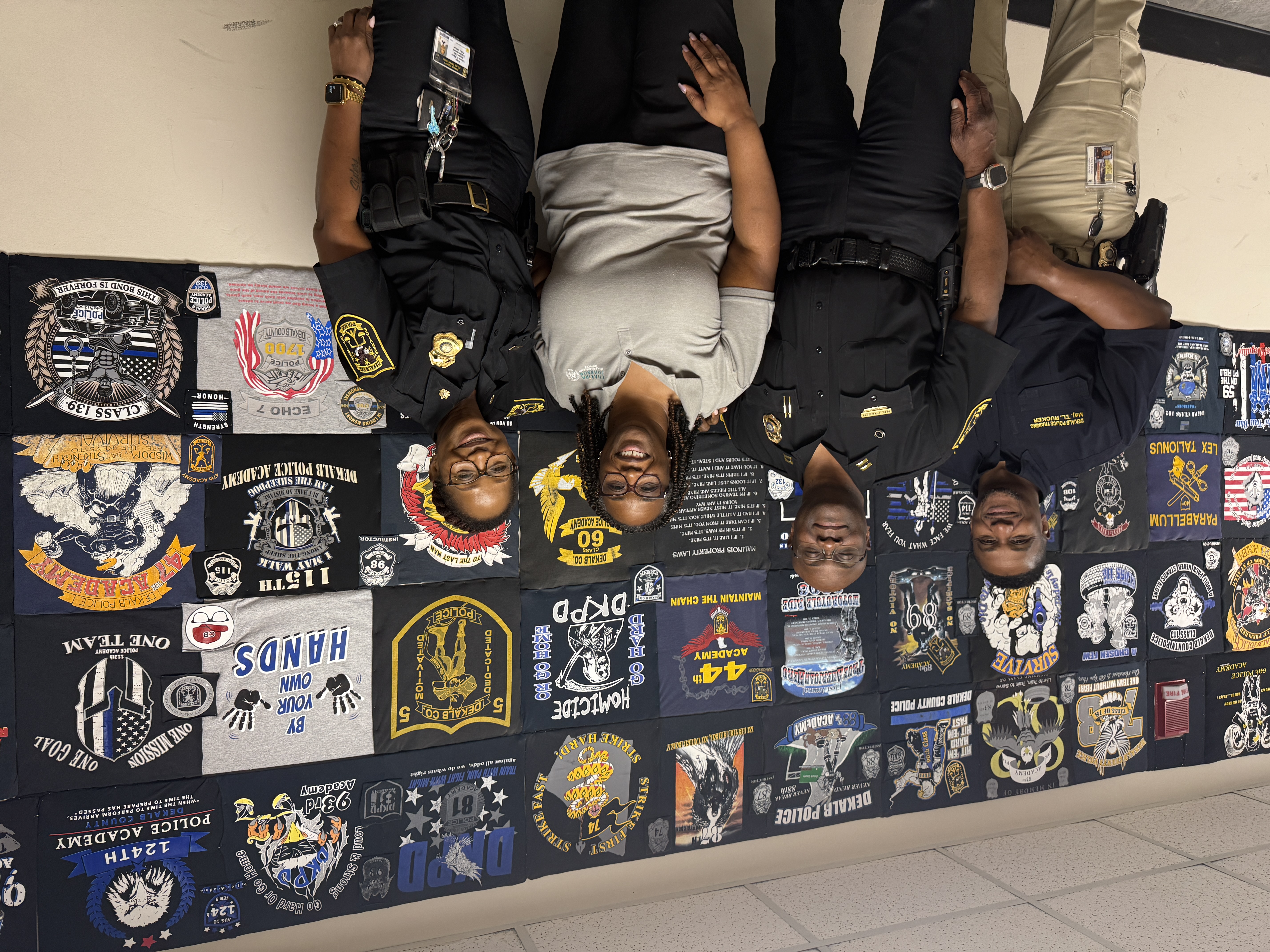 Presiding Officer Commissioner Chakira Johnson smiles with three DeKalb County Police personnel during a visit to the East Precinct. Behind them, a large wall is covered with colorful T-shirts created by graduating police academy classes, featuring distinctive class logos, artwork and mottos.