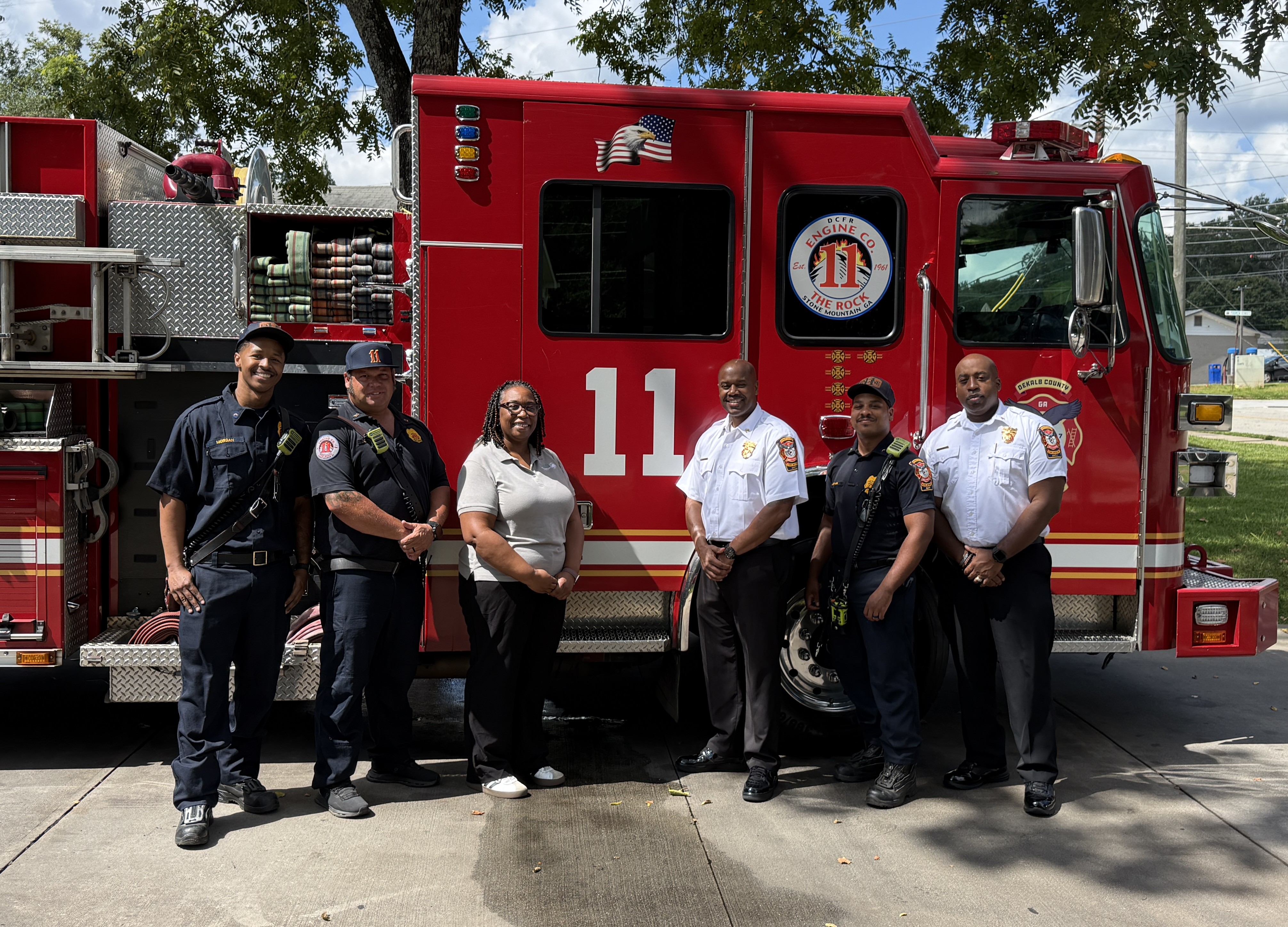Presiding Officer Commissioner Chakira Johnson stands with five DeKalb County Fire Rescue personnel in front of bright red Engine 11 during a visit to Fire Station 11. The group poses outdoors beside the truck, with firefighting equipment and hoses visible along its side.