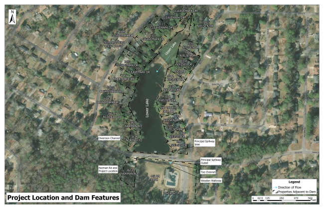 Aerial project map showing the Norman Road Dam and Lower Lake, surrounding residential properties, nearby streets and key dam features. Labels identify the diversion channel, principal spillway inlet and outlet, toe channel, wooden walkway, Norman Road project location and the direction of water flow.