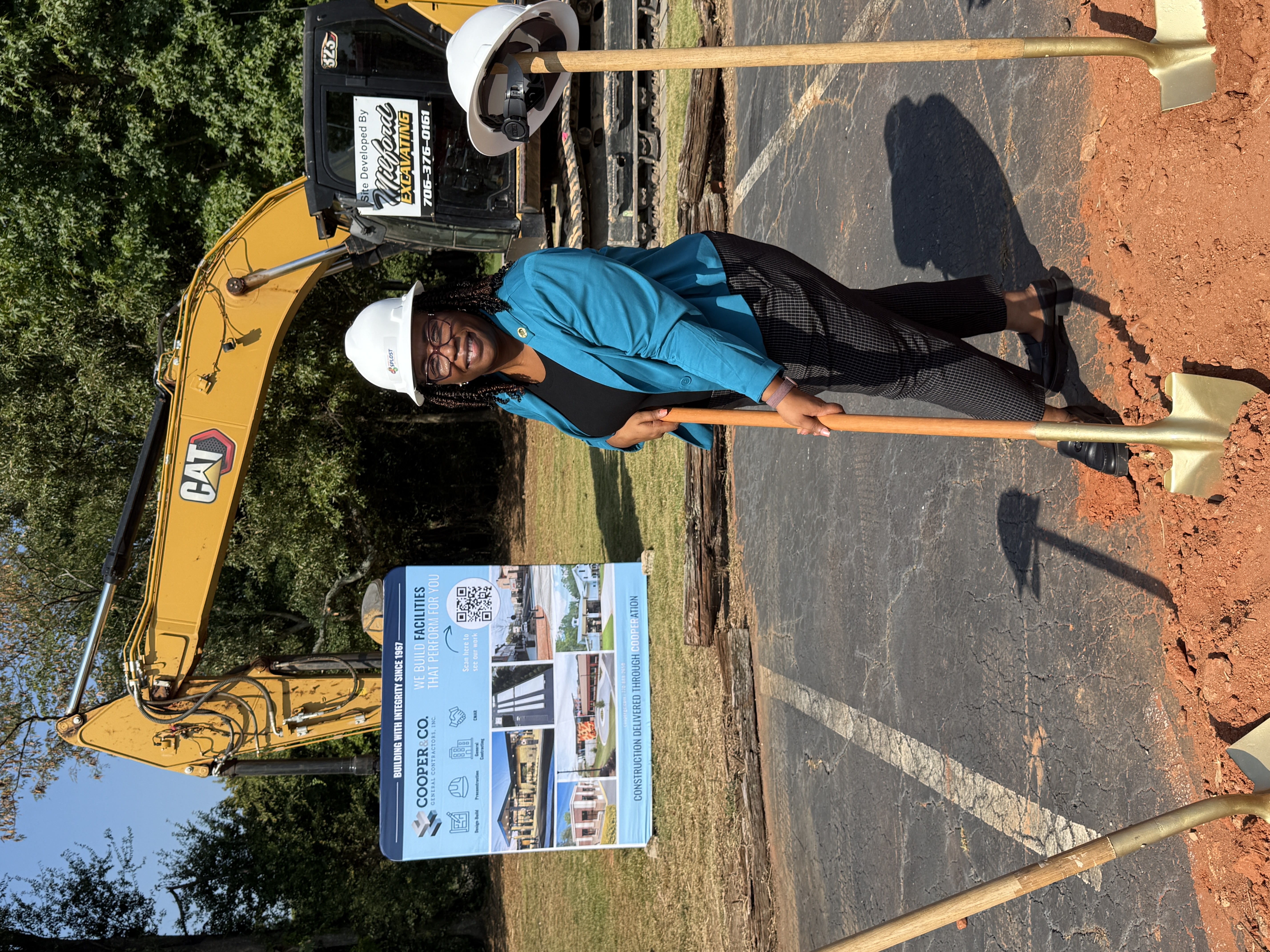 Presiding Officer Commissioner Chakira Johnson, wearing a white SPLOST hard hat, smiles while holding a gold ceremonial shovel at the Fire Station No. 5 groundbreaking. Construction equipment and project signage are visible behind her.