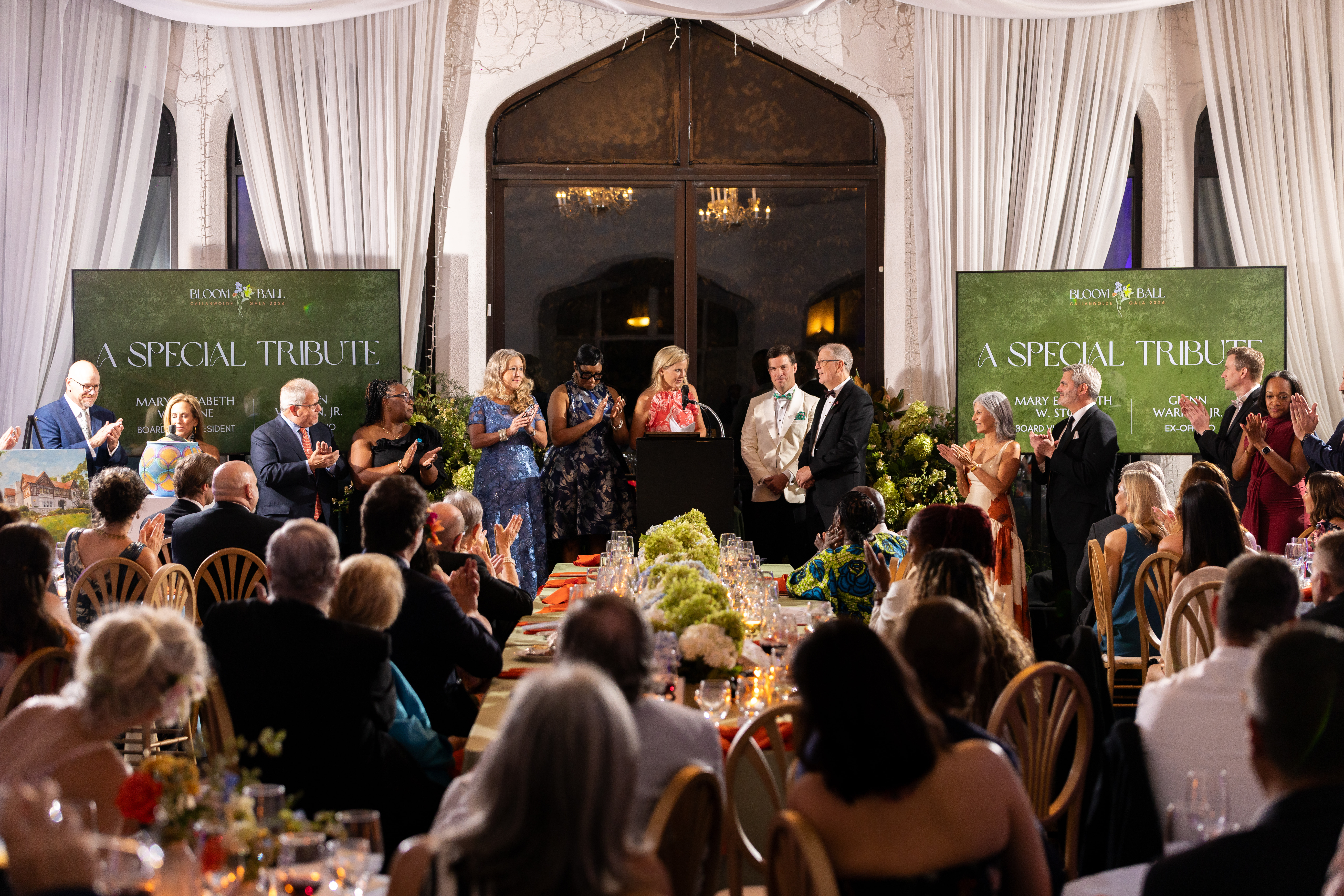 A wide view of Callanwolde Fine Arts Center’s Bloom Ball shows guests seated at decorated dinner tables while people stand and applaud around a podium at the front of the room. Two large screens display “A Special Tribute,” and the elegant ballroom is framed by tall draped windows, floral arrangements and candlelit tables.