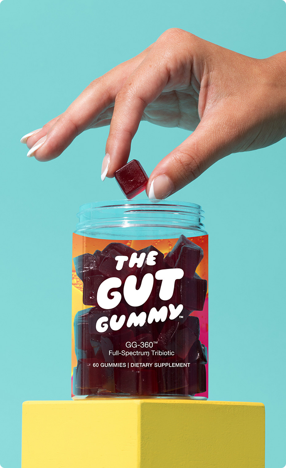 Hand holding a Gut Gummy cube above an open jar with label “The Gut Gummy™ – GG-360™ Full-Spectrum Tribiotic, 60 Gummies” against a teal background.