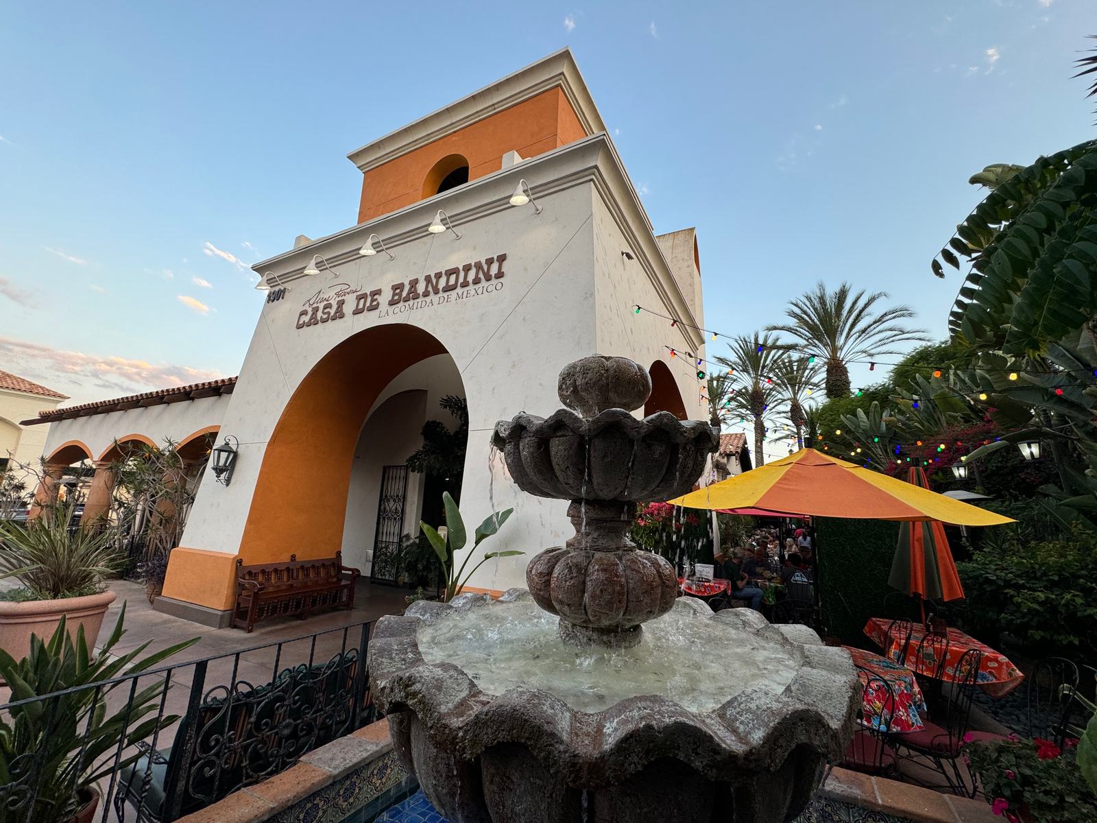 Casa de Bandini's white bell-tower arch and tiered stone fountain at dusk, with patio umbrellas and palms, The Forum, Carlsbad