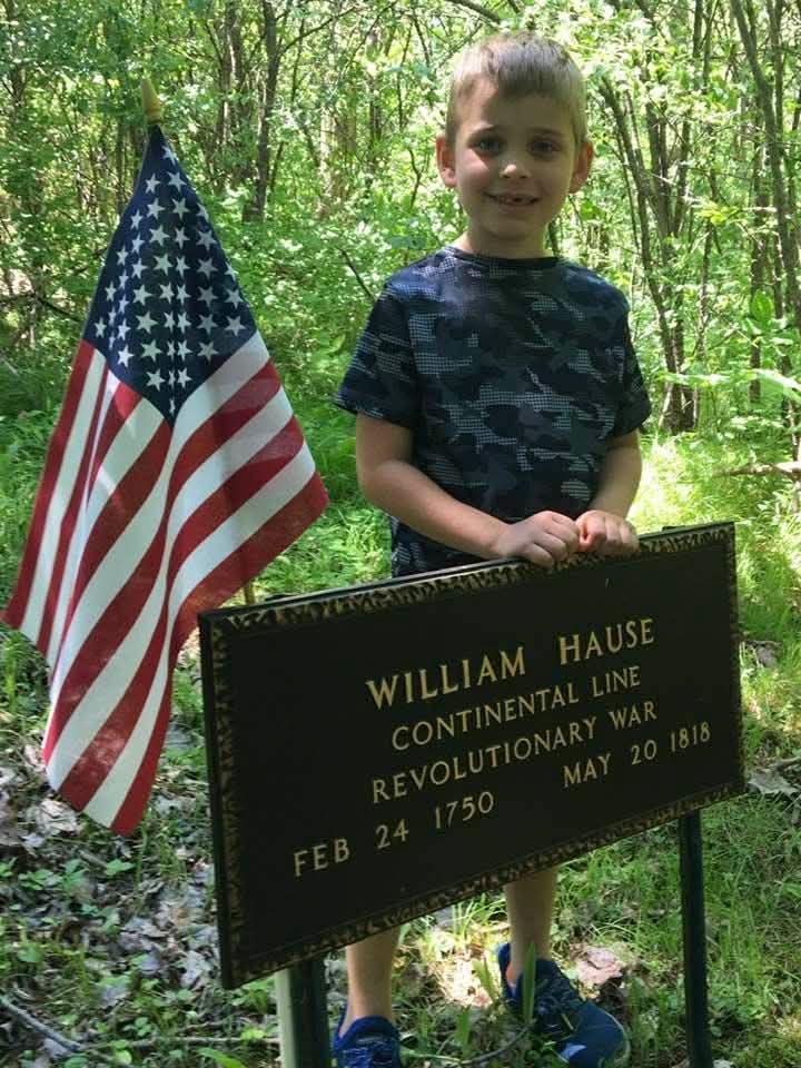 Young boy stands beside the grave marker of Revolutionary War Continental Line veteran William Hause, holding an American flag at his burial site in Tyrone, New York.